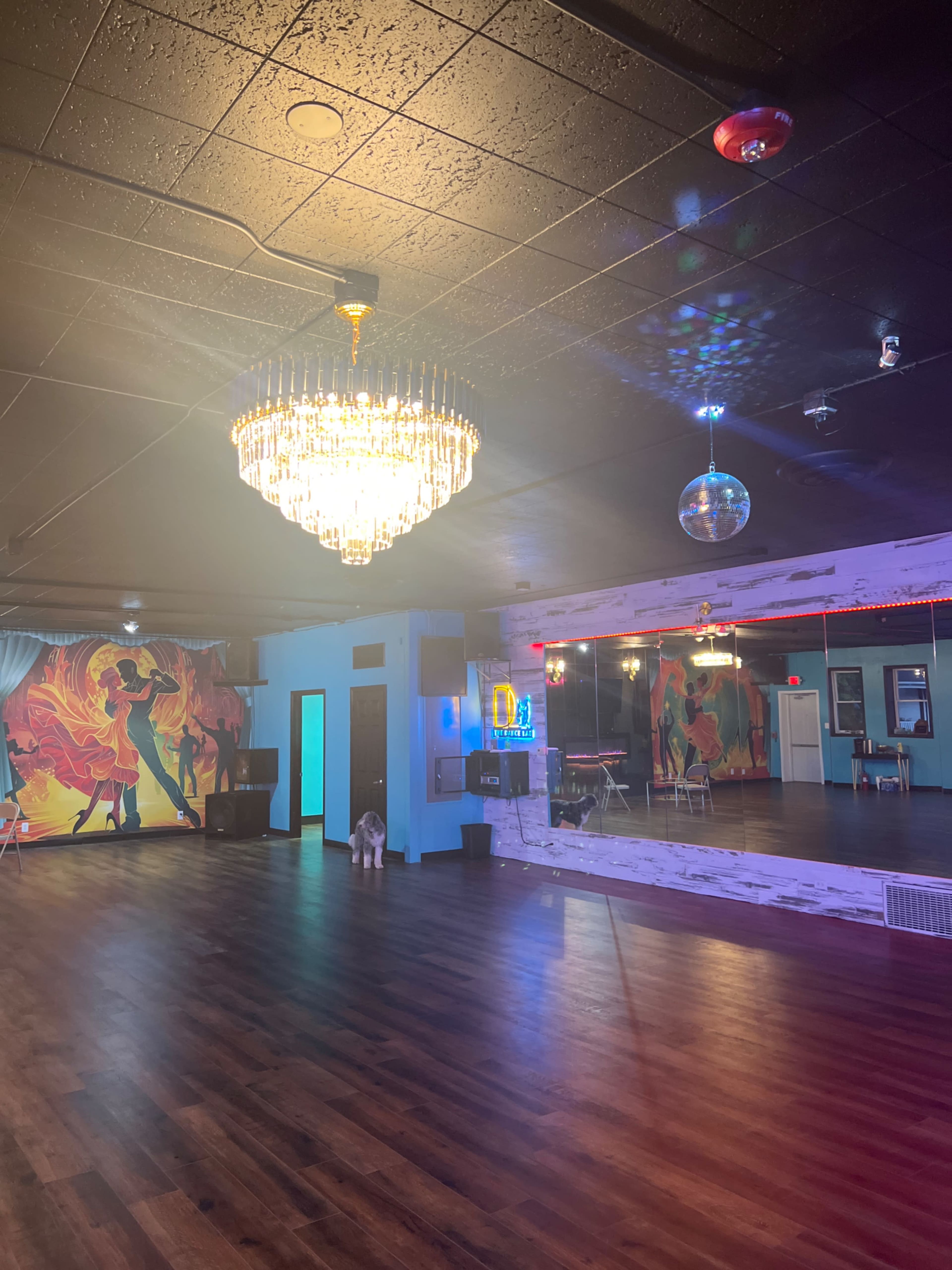 A spacious dance studio featuring a chandelier, mirrored wall, colorful mural, and wooden flooring.