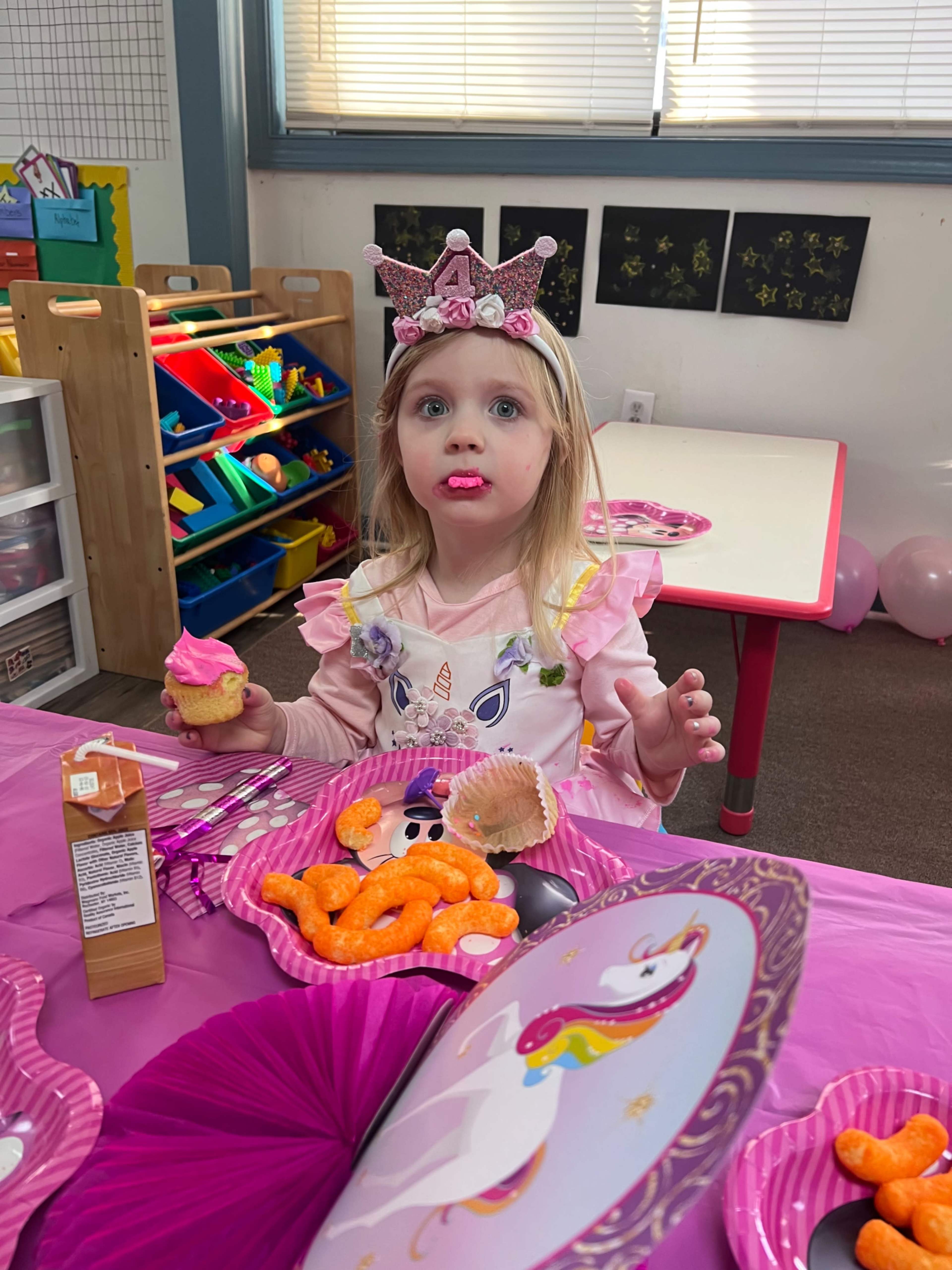 A young child wearing a crown sits at a table decorated for a party, holding a cupcake and surrounded by colorful snacks on pink plates.