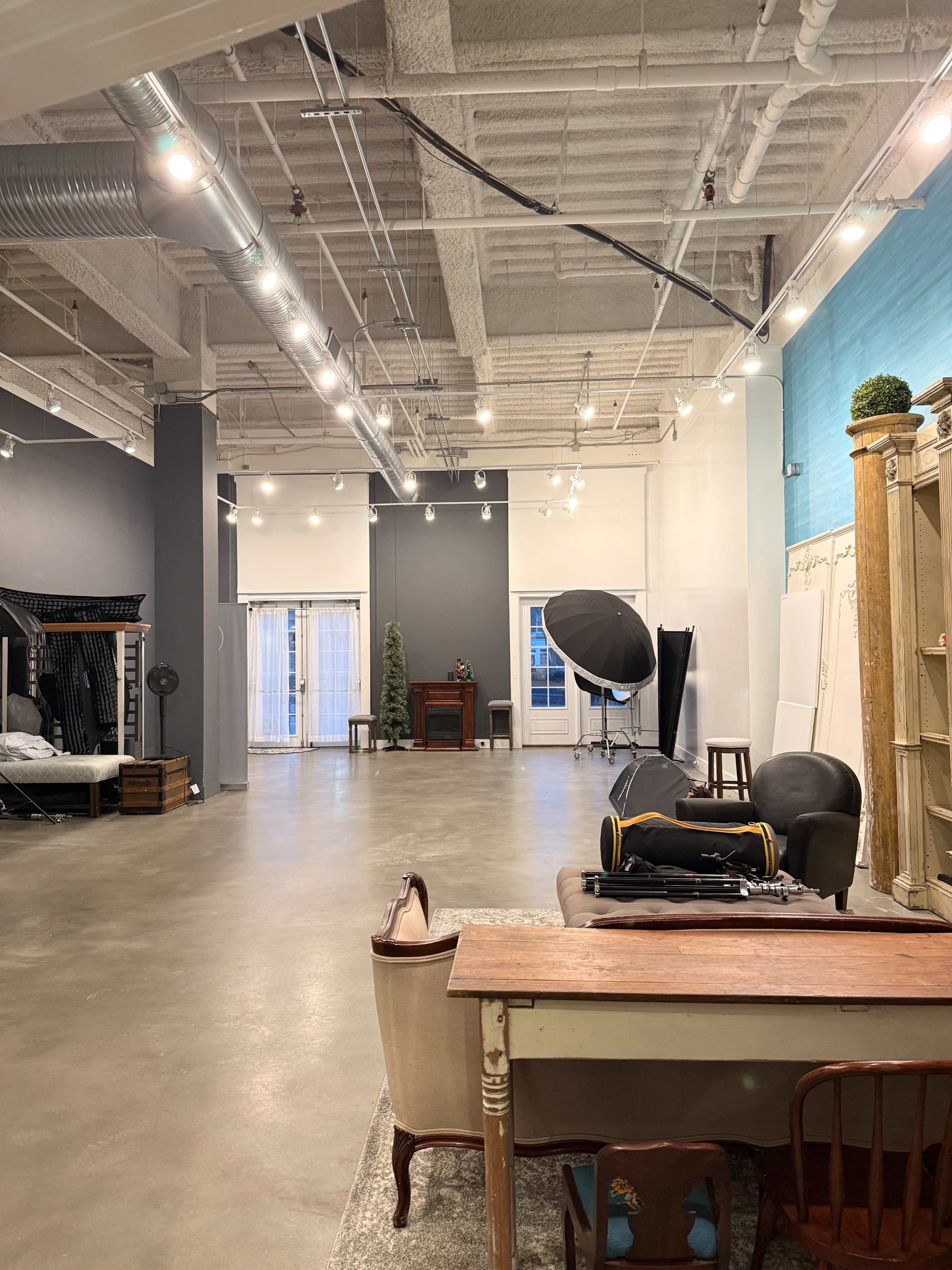 The image shows a spacious, well-lit studio with gray and blue walls, featuring various furniture pieces and lighting equipment arranged throughout the room.