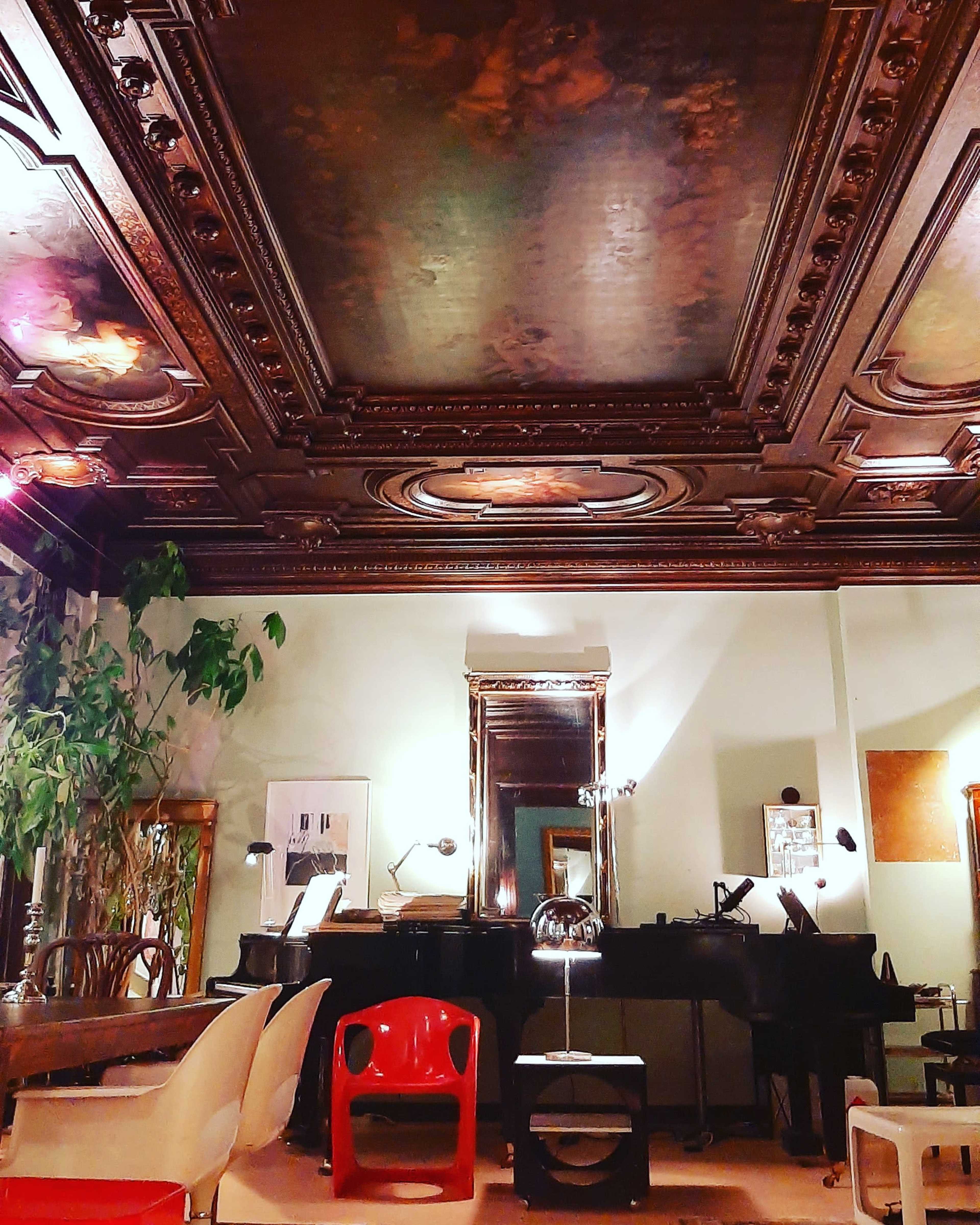 A room with a decorative wooden ceiling, a black piano, various chairs, and a large mirror reflecting the space.