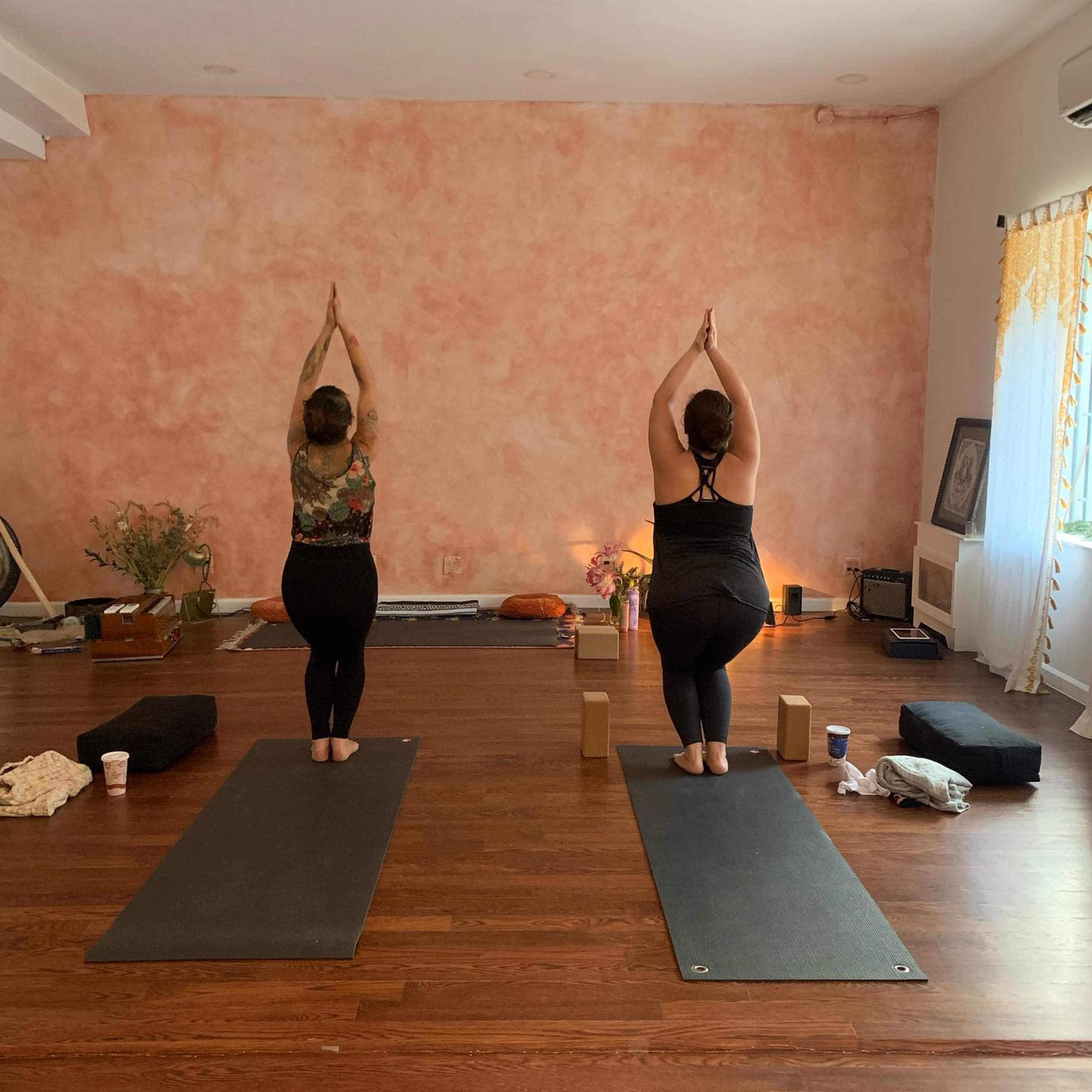 Two individuals practice yoga with their hands raised in a studio space that features wooden floors and a soft pink wall.