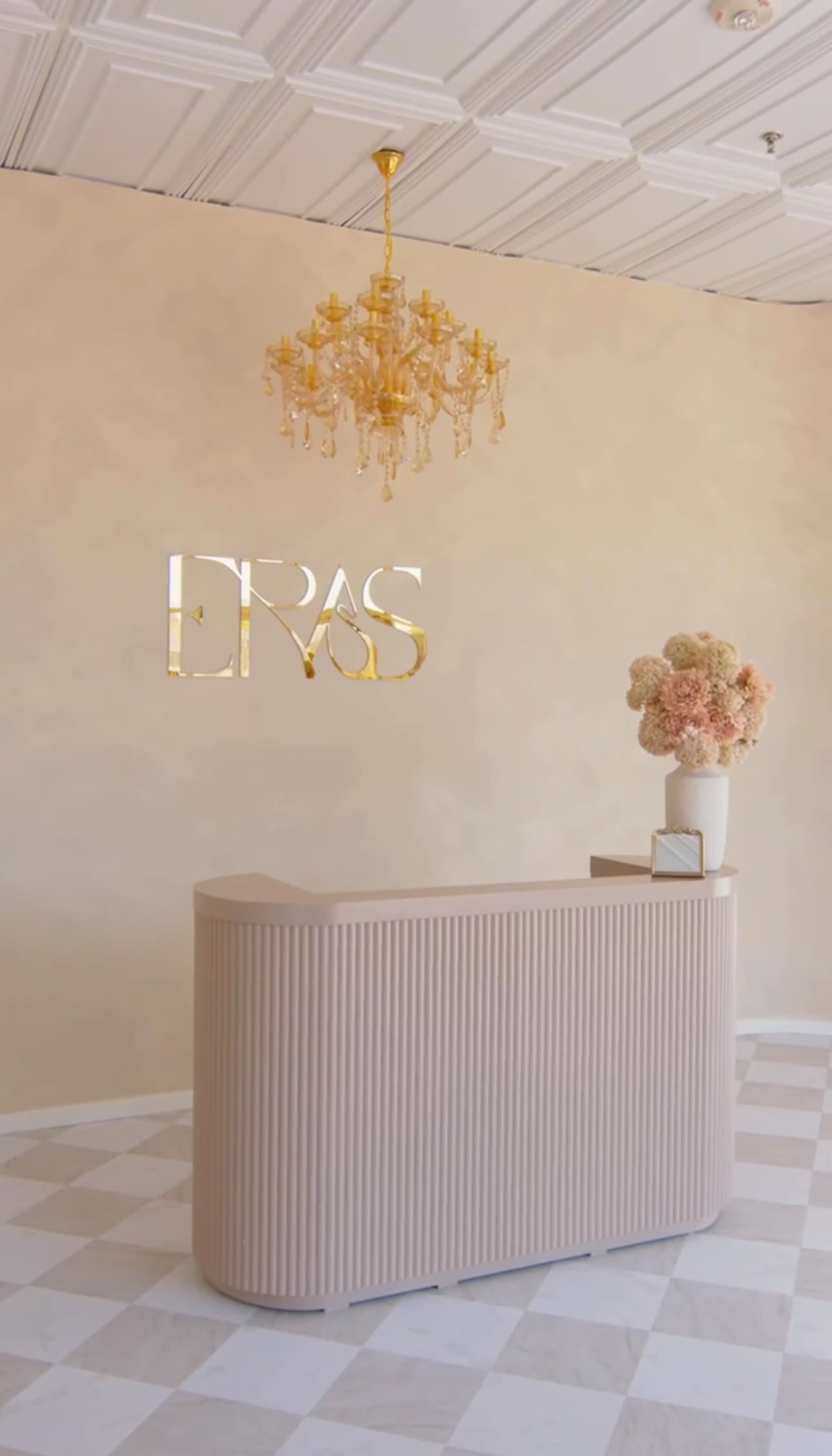 The image depicts a modern reception area with a curved desk, a floral arrangement in a vase, and an elegant chandelier hanging from the ceiling.