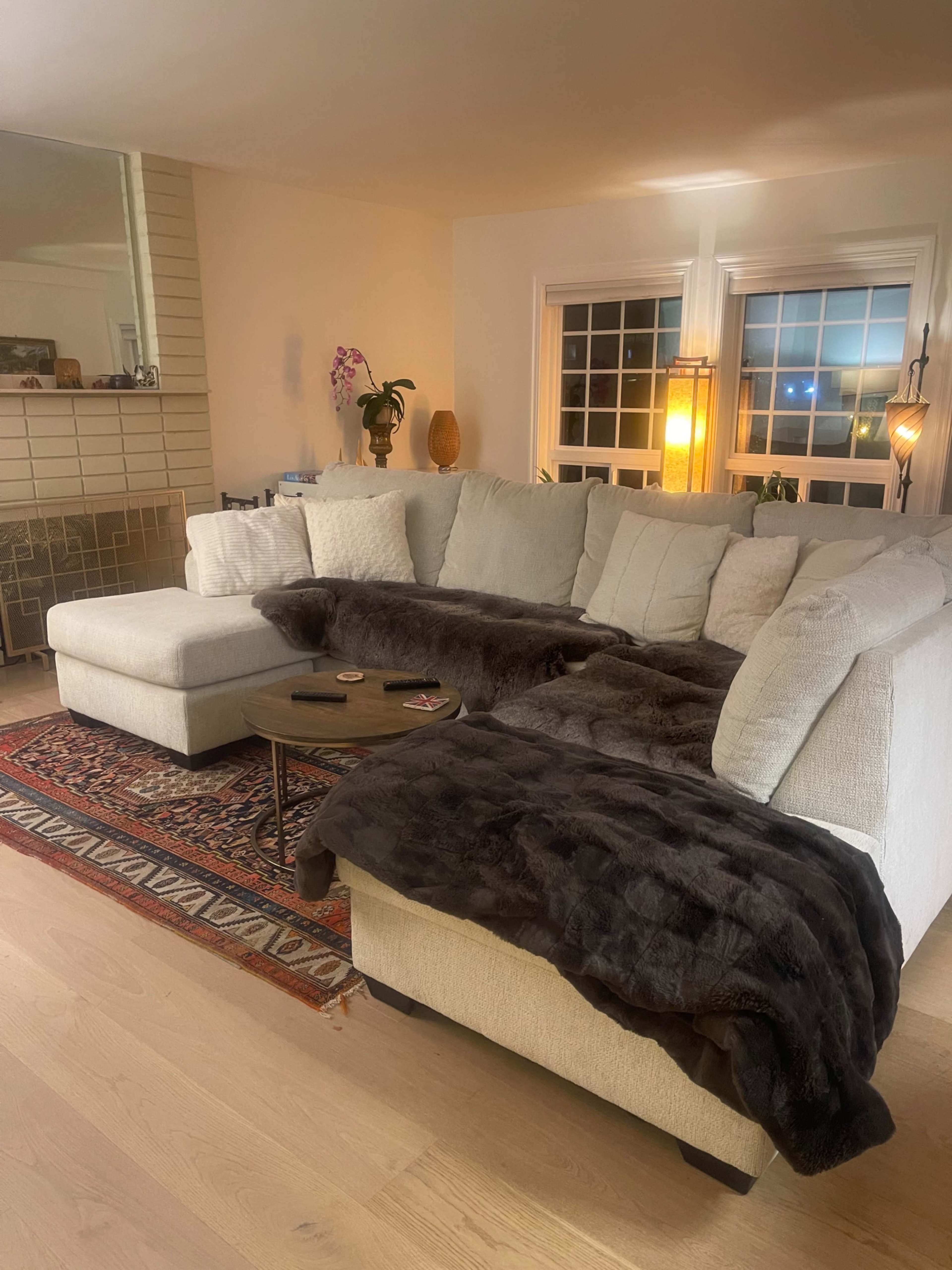 The image shows a cozy living room featuring a large L-shaped sofa with a fur throw, a wooden coffee table, and a patterned area rug, accompanied by decorative plants and warm lighting.