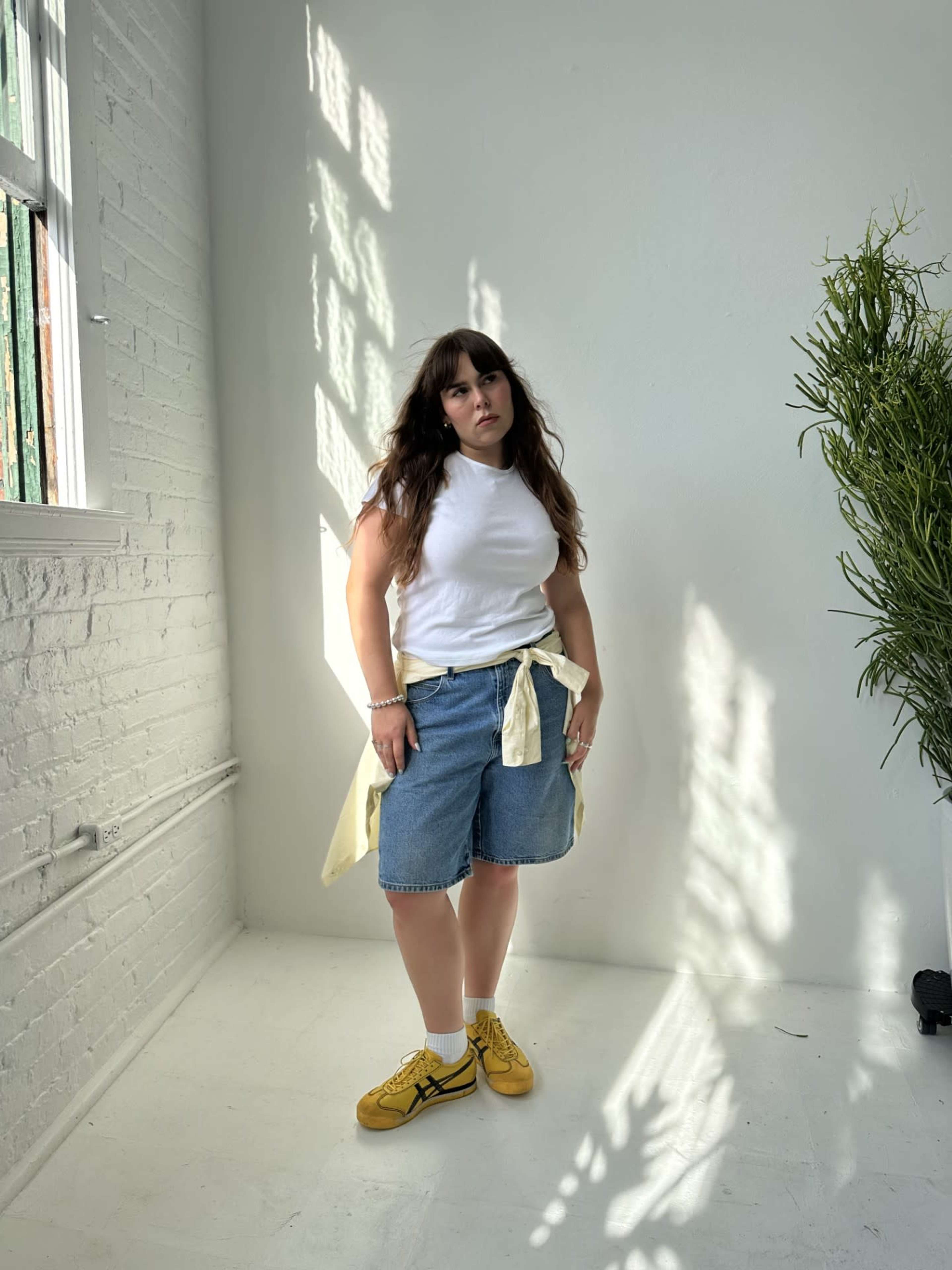 A person stands in a brightly lit room with white walls and a large plant, dressed in a white top, denim shorts, and yellow shoes.