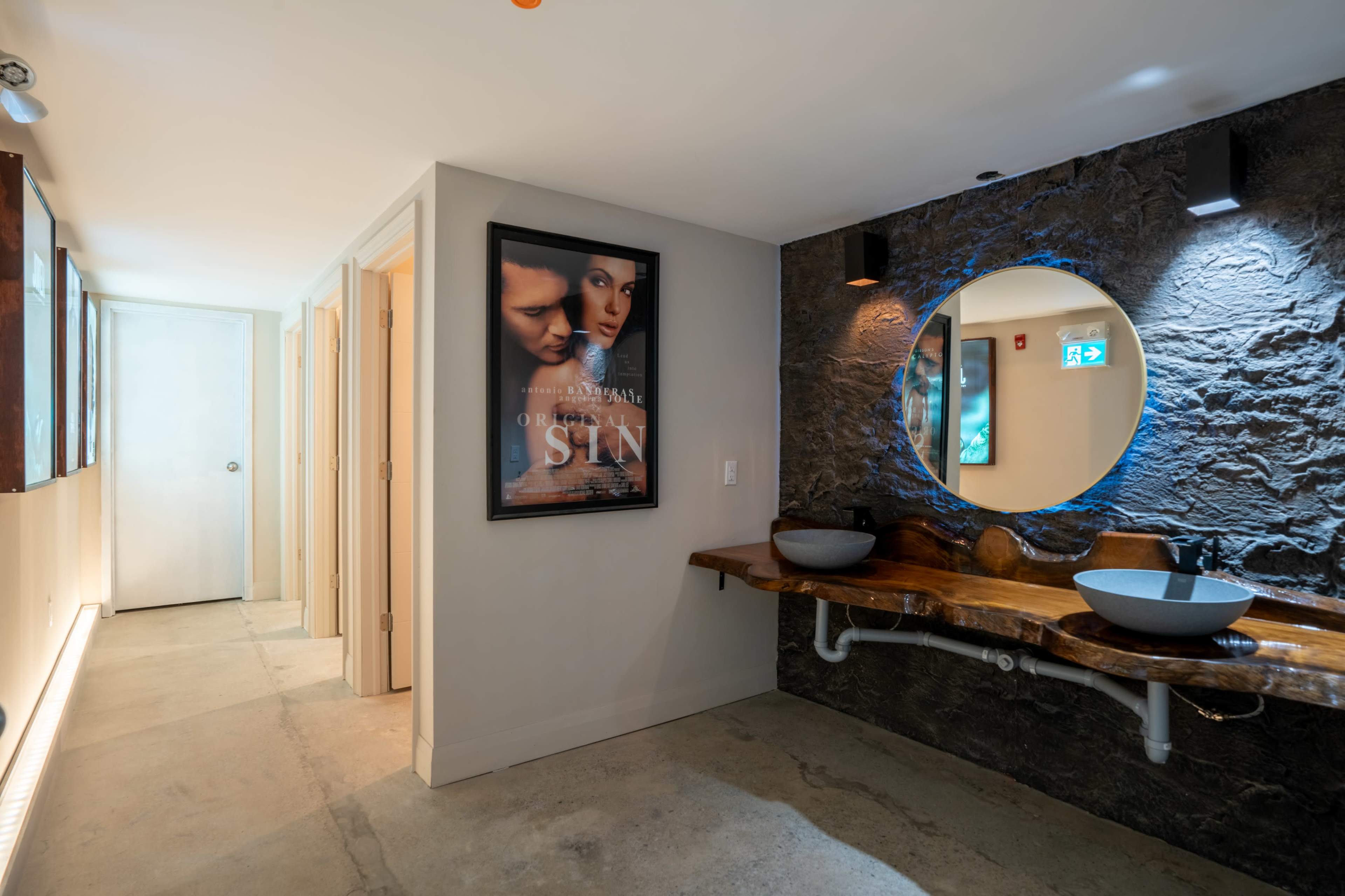A modern bathroom with a stone wall, wooden sink counter, circular mirror, and a long hallway leading to a door, illuminated by soft lighting.