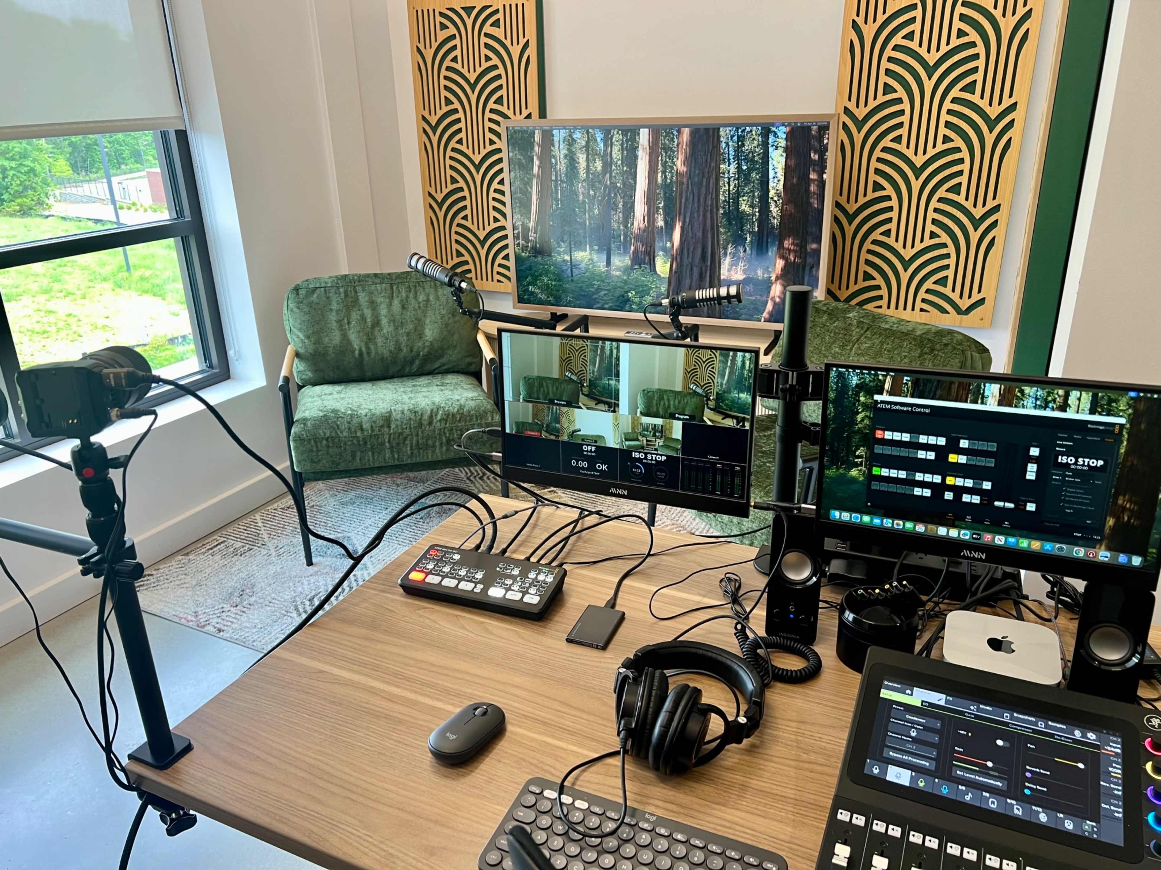 The image shows a modern recording studio setup with multiple computer monitors, a microphone, a camera on a tripod, and a cozy green couch in a well-lit room featuring nature-themed decor.