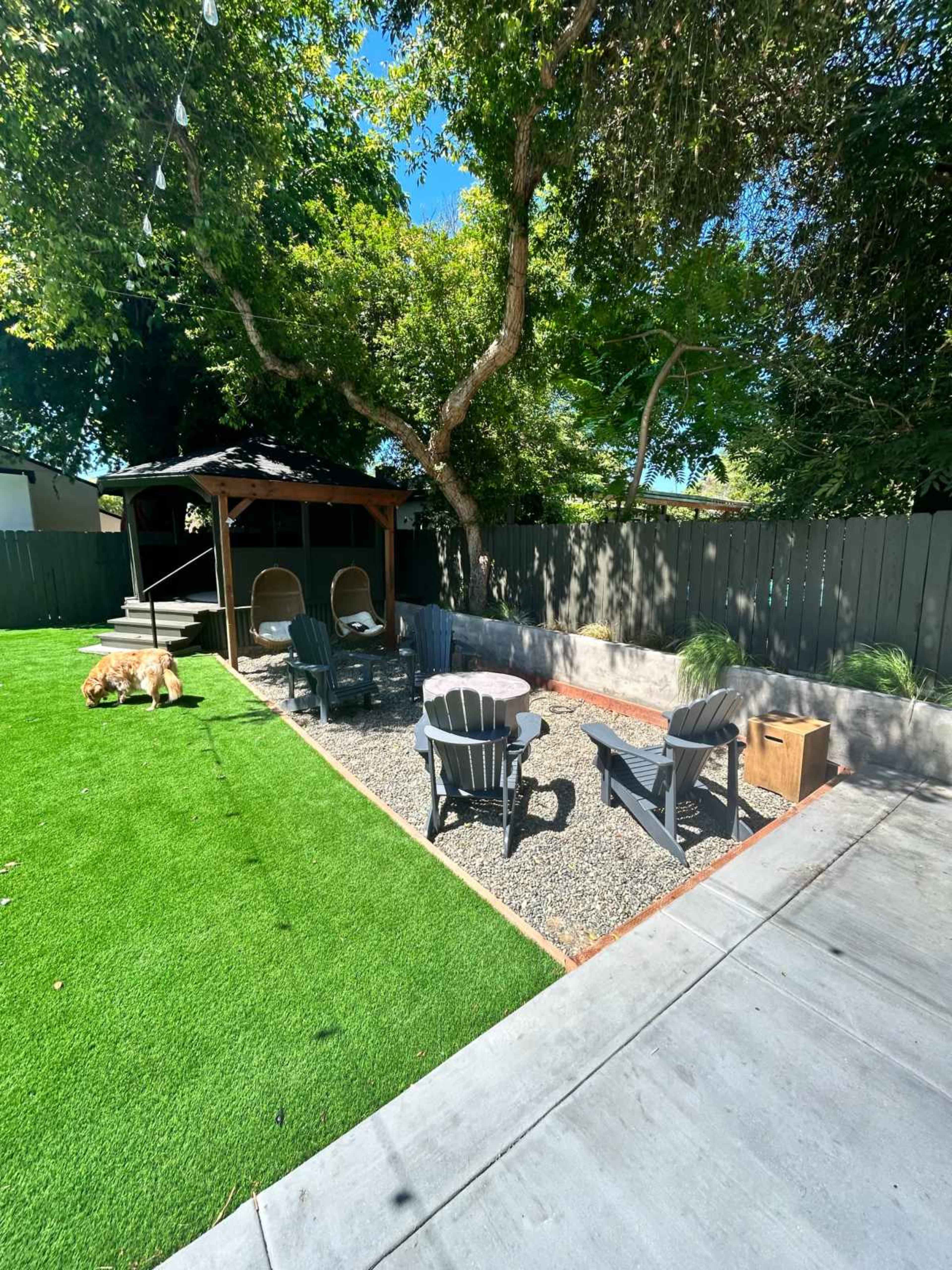The image shows a backyard featuring a seating area with chairs around a fire pit, surrounded by gravel, artificial grass, and a gazebo in the background.