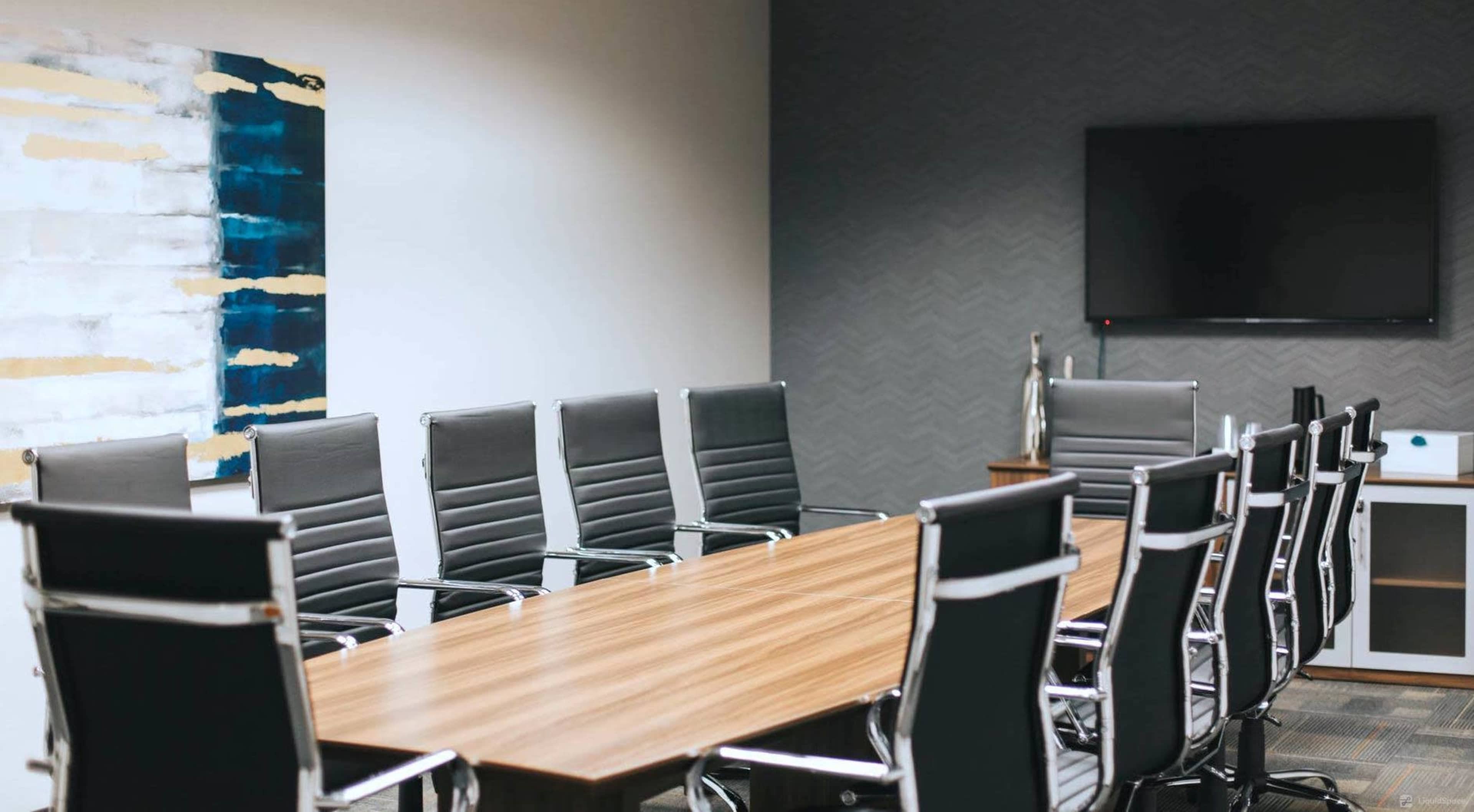 A conference room features a long wooden table surrounded by black leather chairs, with a large wall-mounted television and a piece of abstract art on the wall.