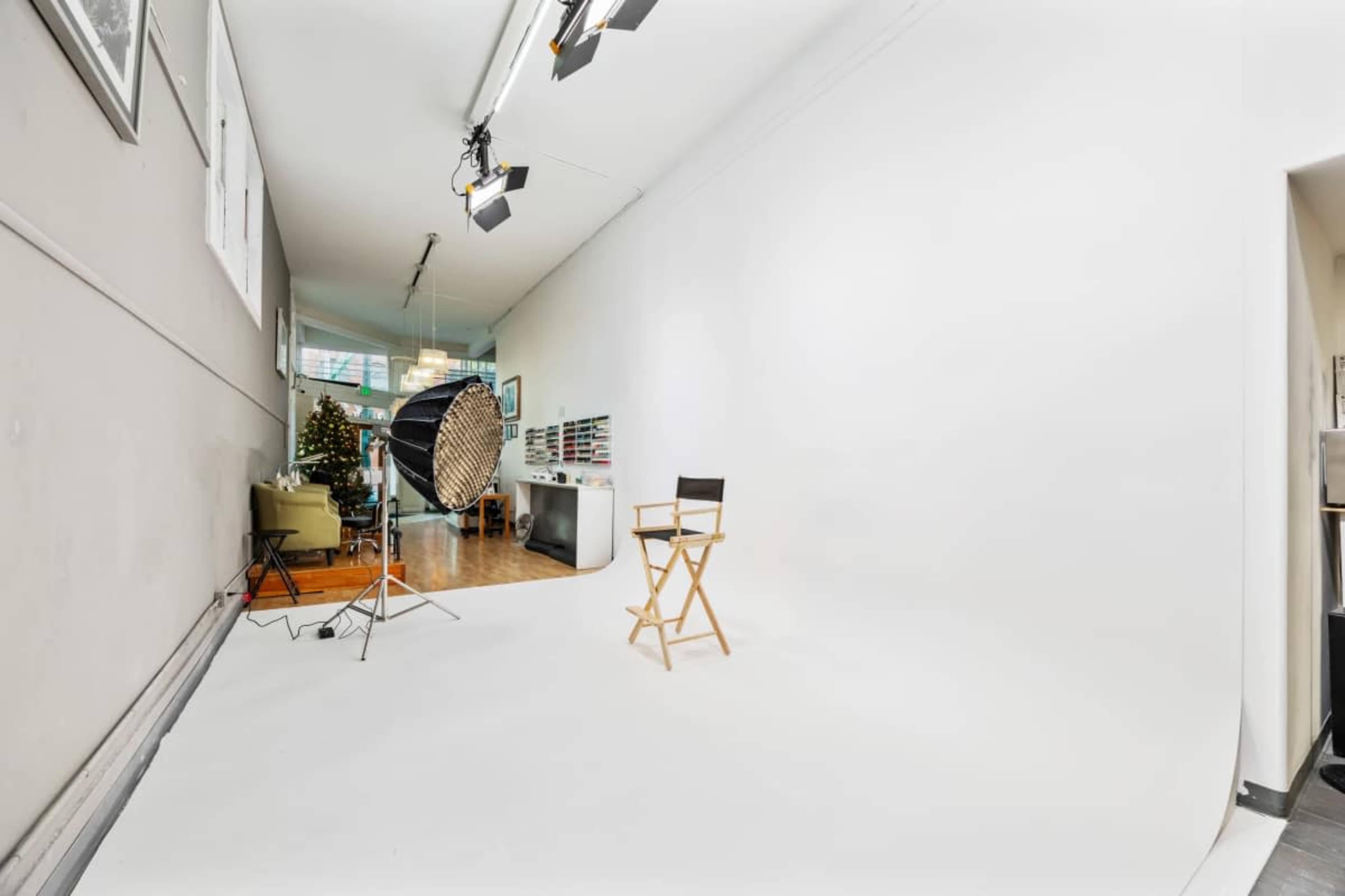The image shows a photo studio with a white backdrop, a director's chair, and a lighting setup, all positioned in an open space that includes a decorative Christmas tree in the background.