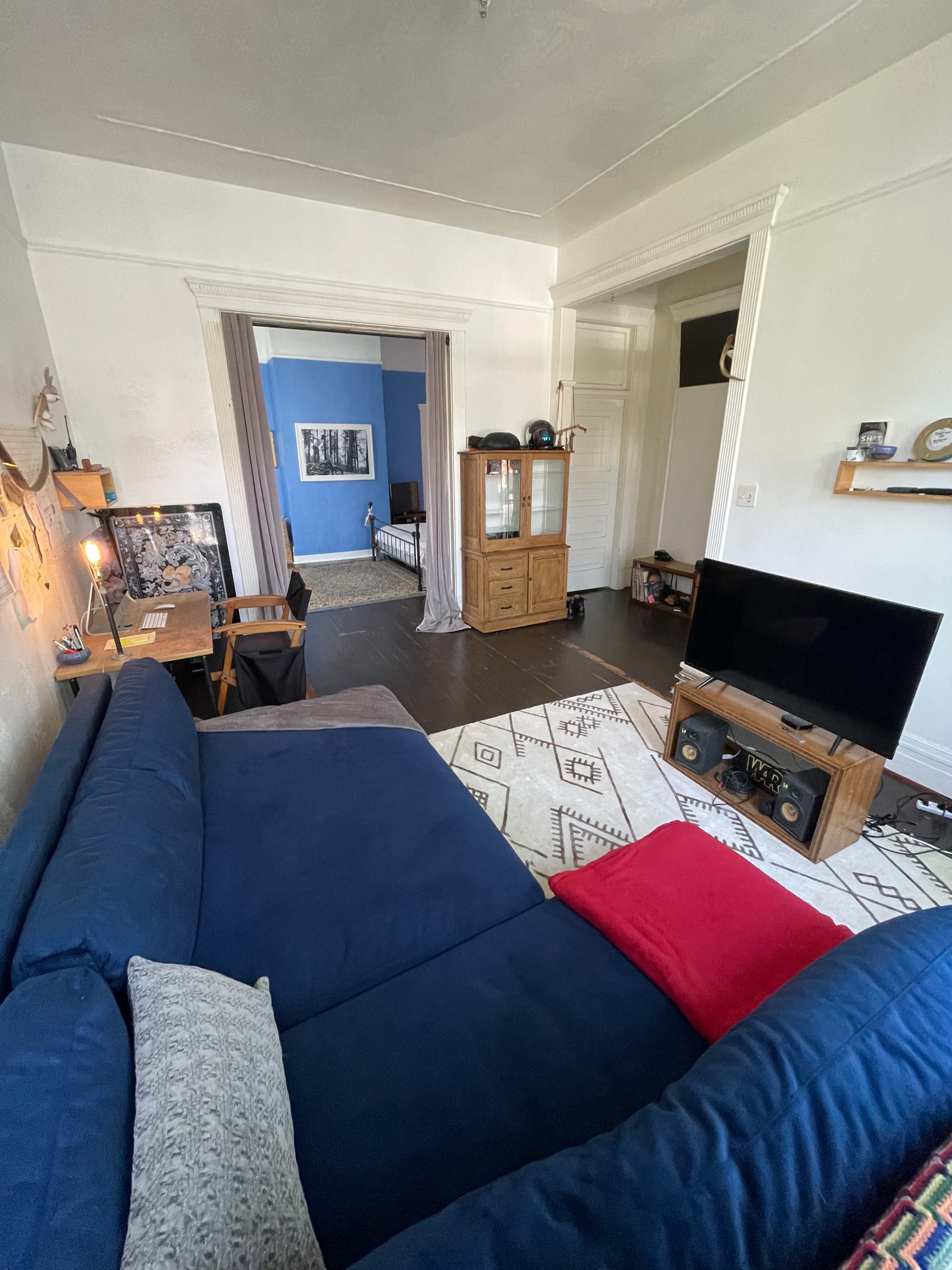 The image shows a living room with a blue couch, a television stand, a small dining area, and a view into an adjacent blue-walled bedroom.