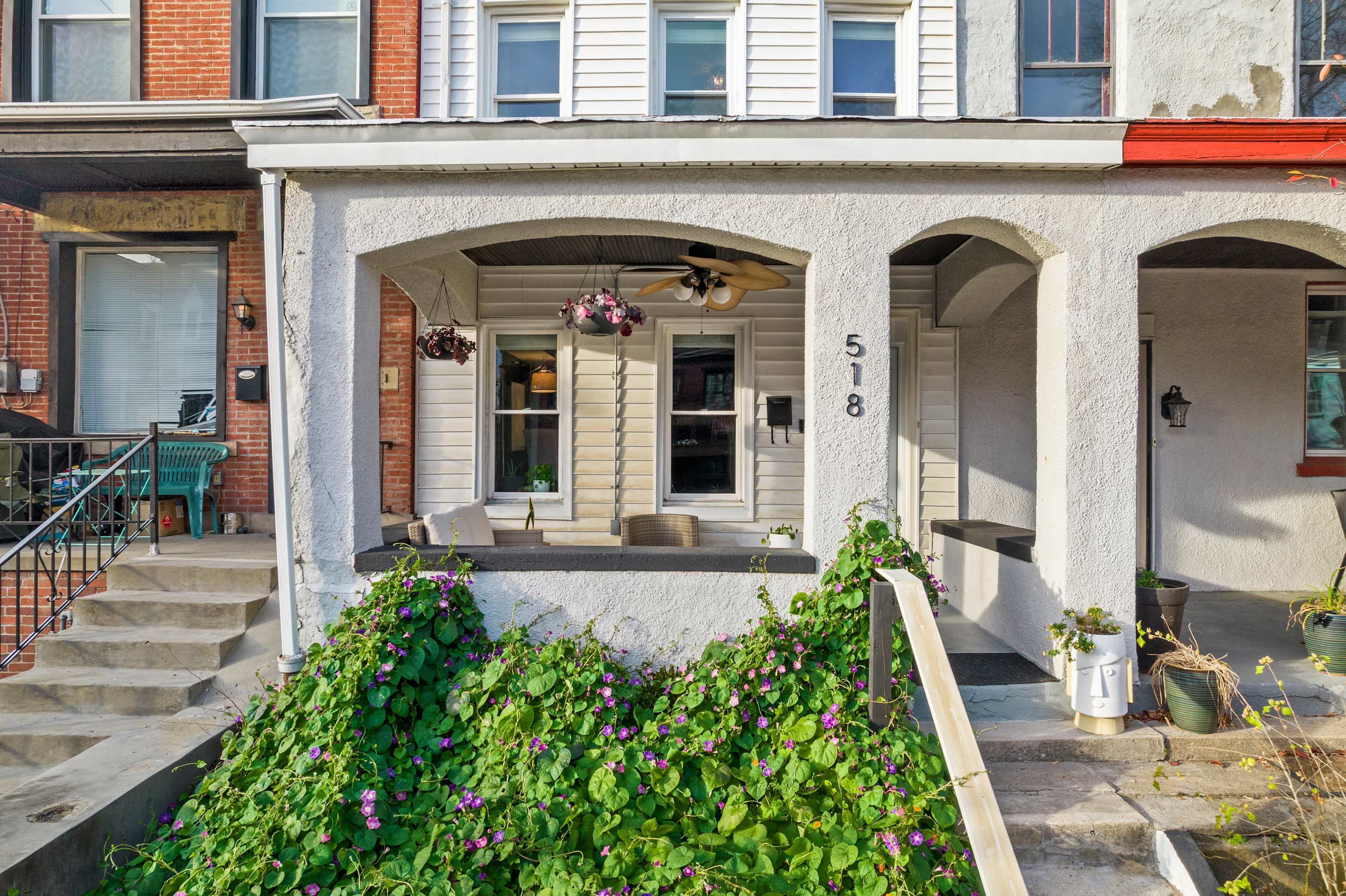 A porch with a white exterior, featuring two chairs and hanging flower pots, in front of a house numbered 518, with brick and wooden elements visible on adjacent homes.