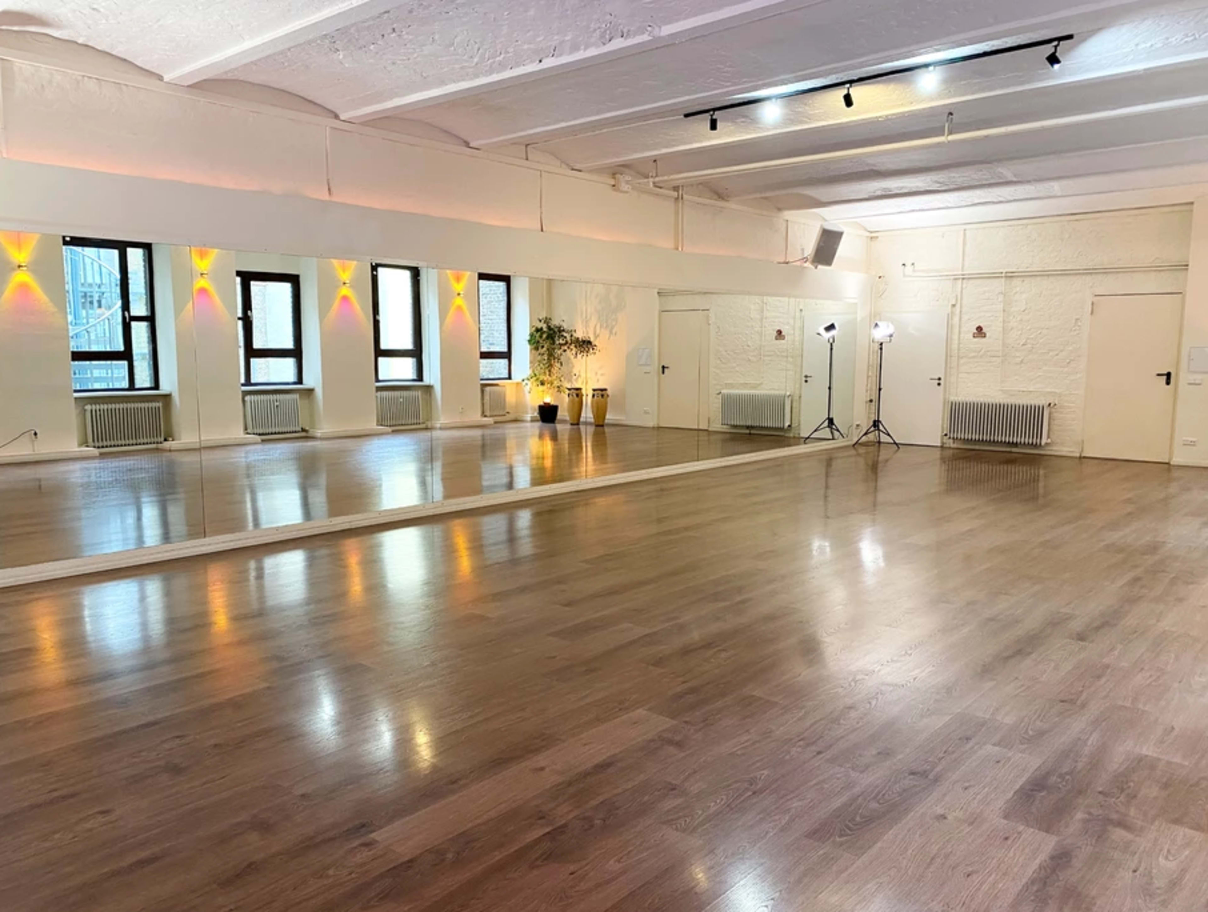 A spacious dance studio with wooden flooring, large mirrors, and natural light streaming through several windows.