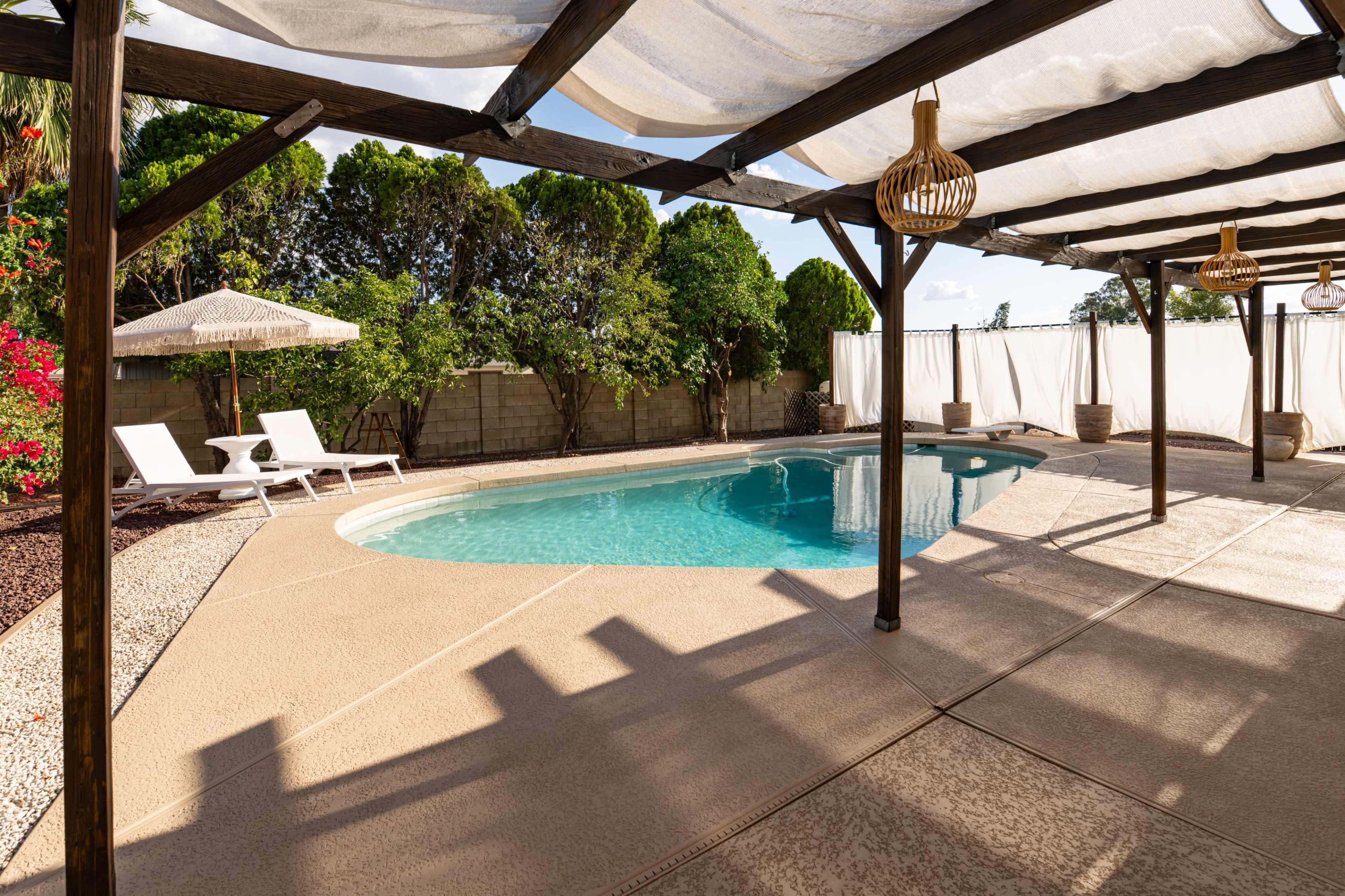 The image shows a swimming pool surrounded by a patio area, with two lounge chairs and a shaded seating space under a pergola.