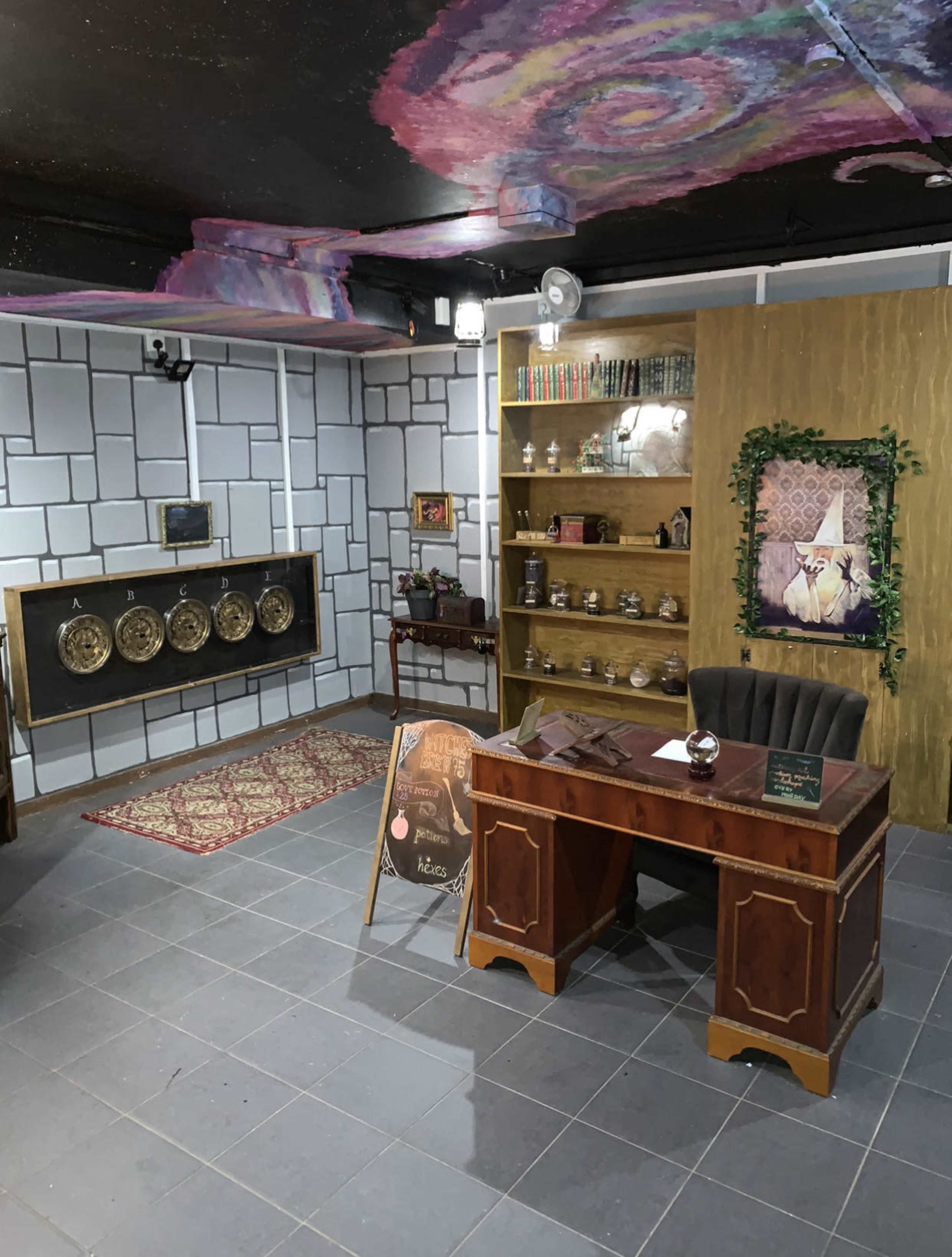 A room with a decorative desk, bookshelves, wall-mounted art, and a patterned rug on a tiled floor.