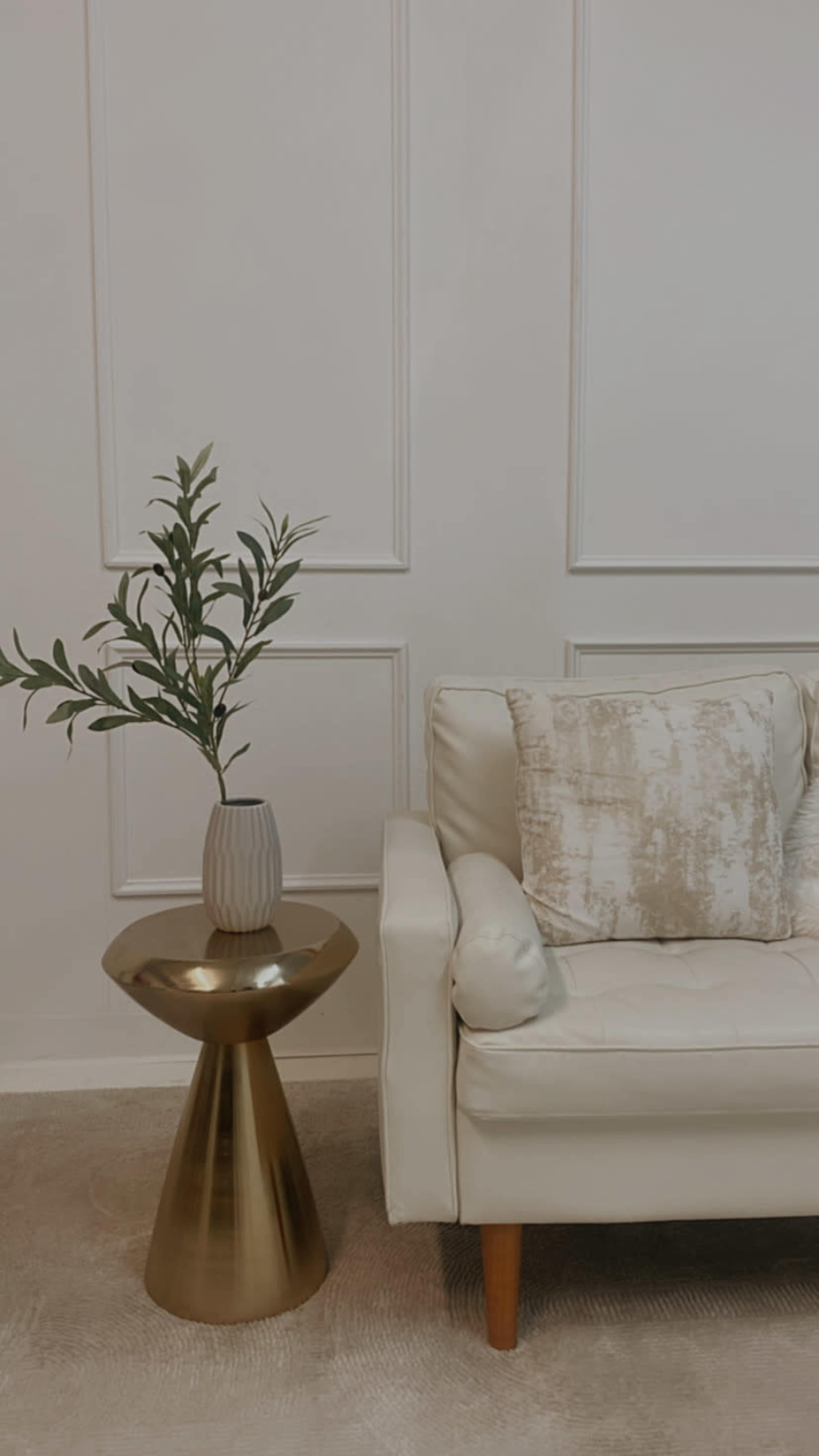 A white sofa sits beside a gold accent table holding a vase with greenery.