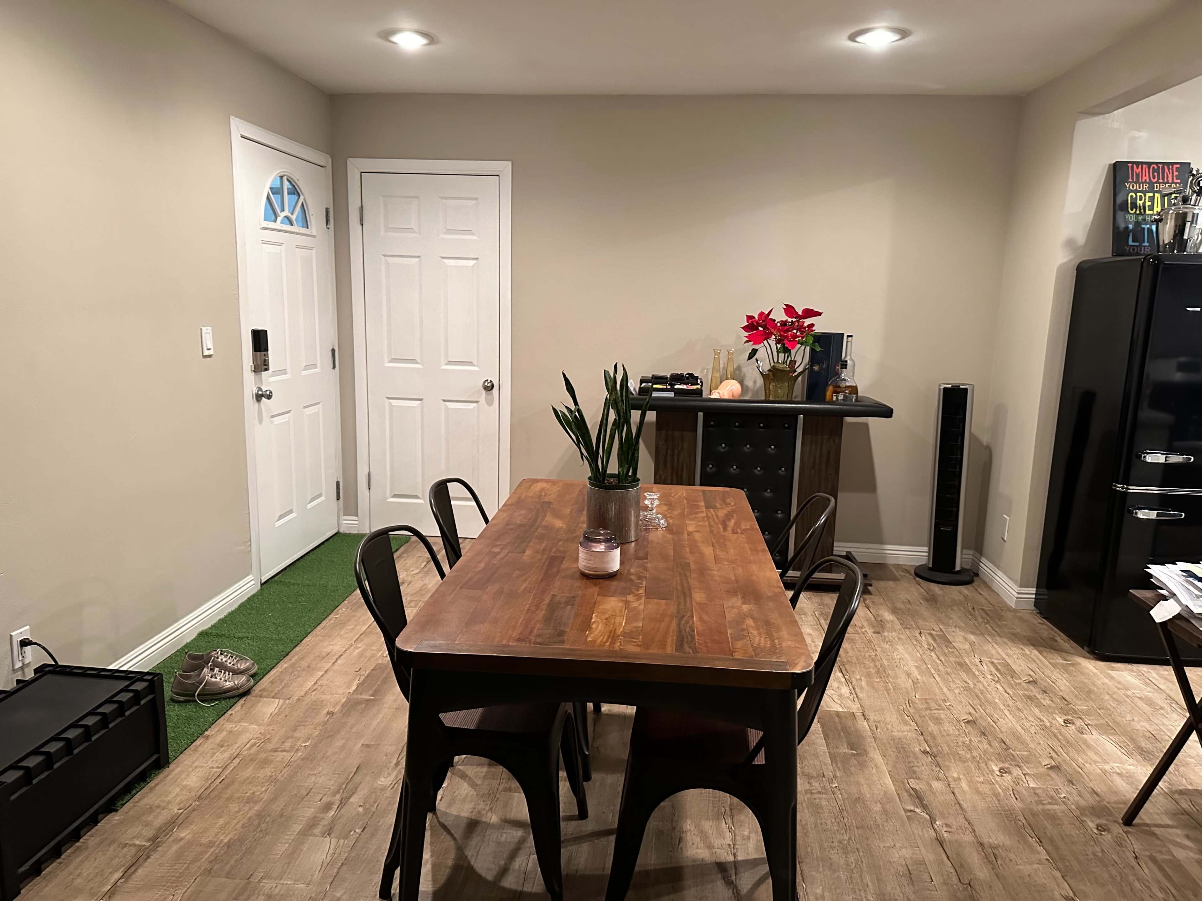 A dining area features a wooden table with metal chairs, a doorway, and a black refrigerator against a light-colored wall.