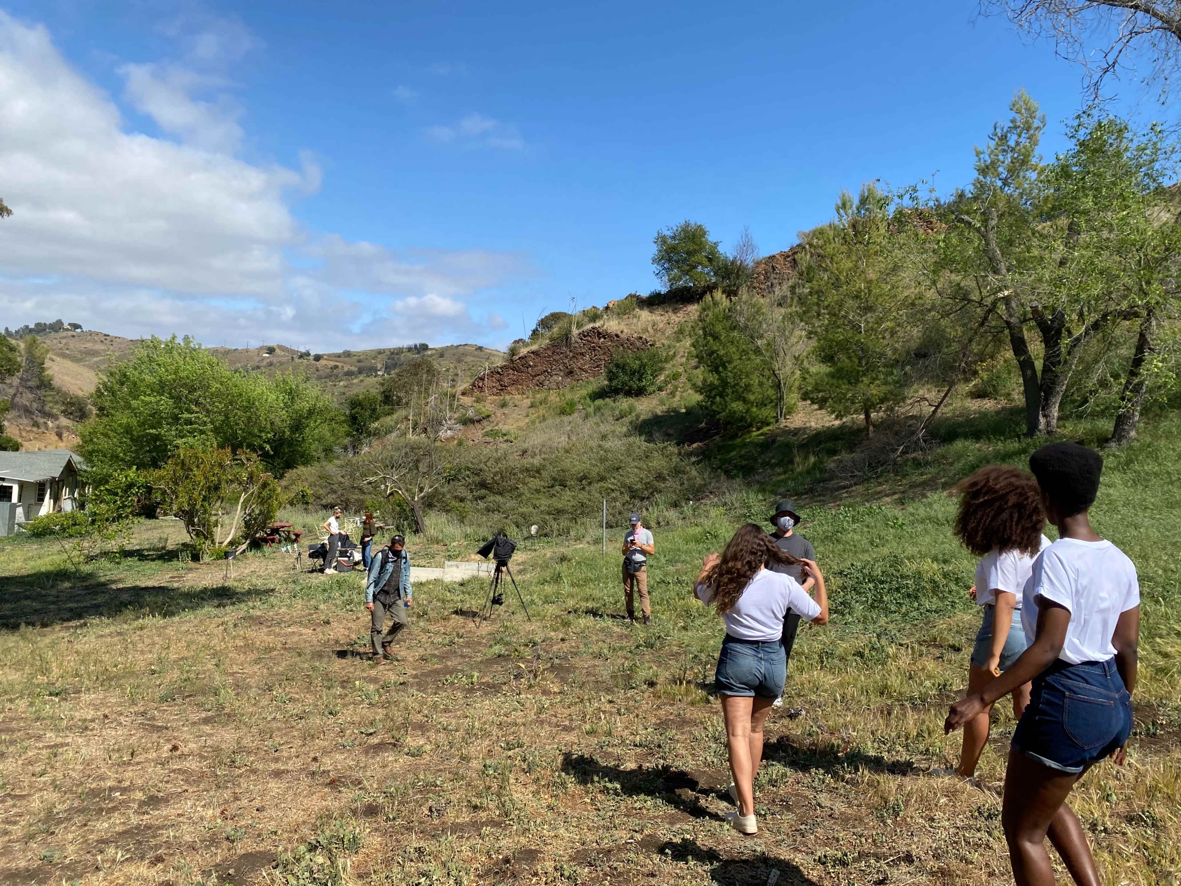 A group of people is walking across a grassy field with trees and hills in the background.