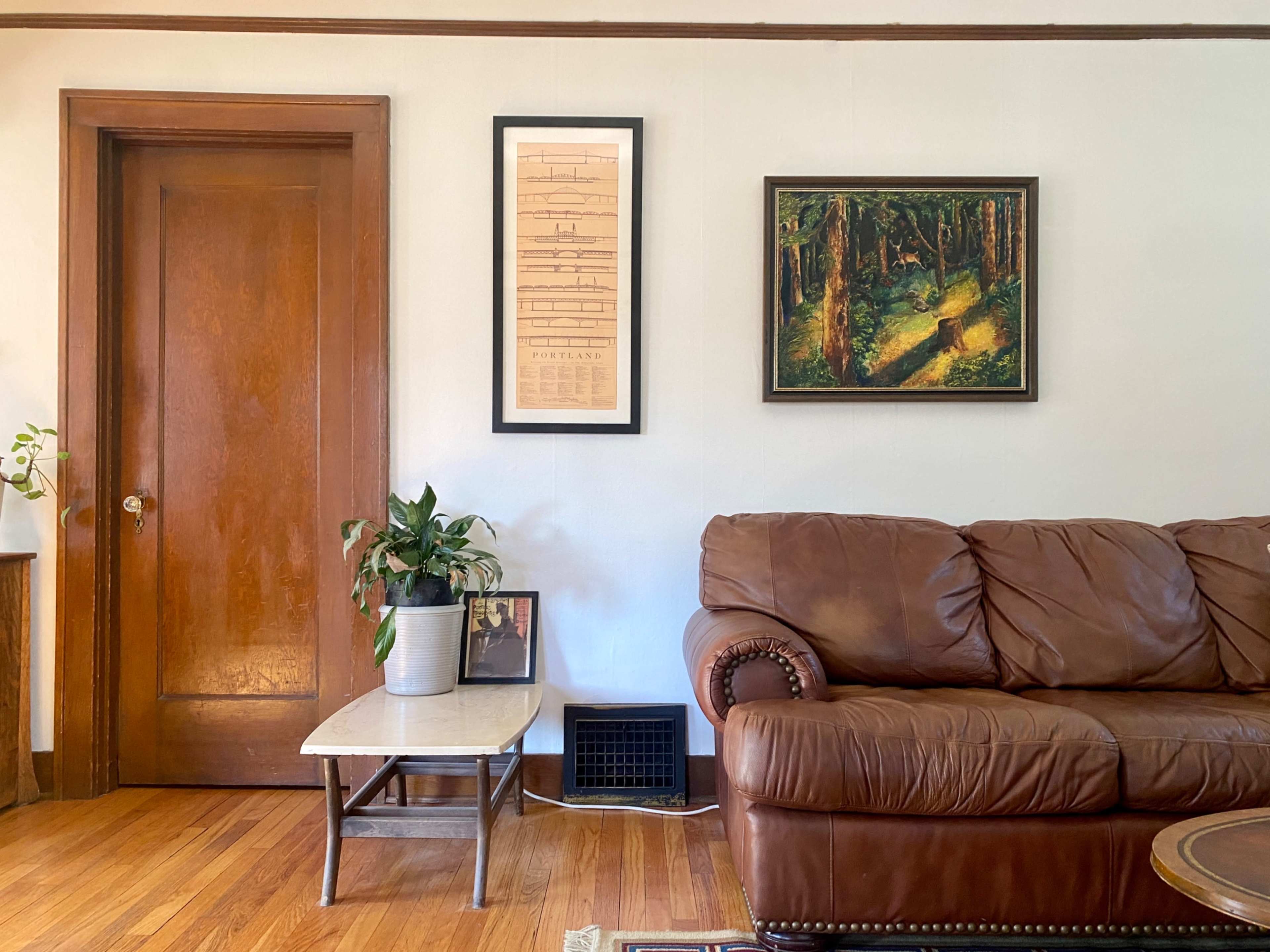 A brown leather couch is positioned next to a wooden door, with a potted plant and framed pictures nearby, and artwork hanging on the wall.