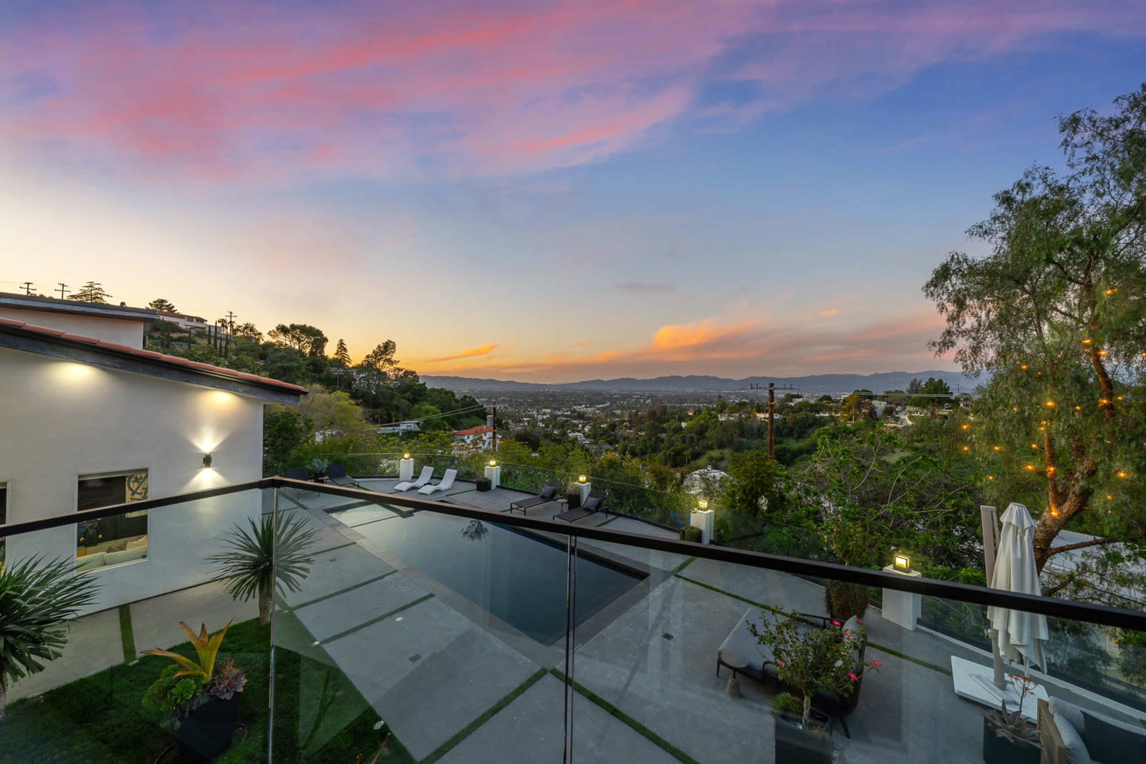 A modern house with a glass railing overlooking a swimming pool and scenic views of a valley at sunset.