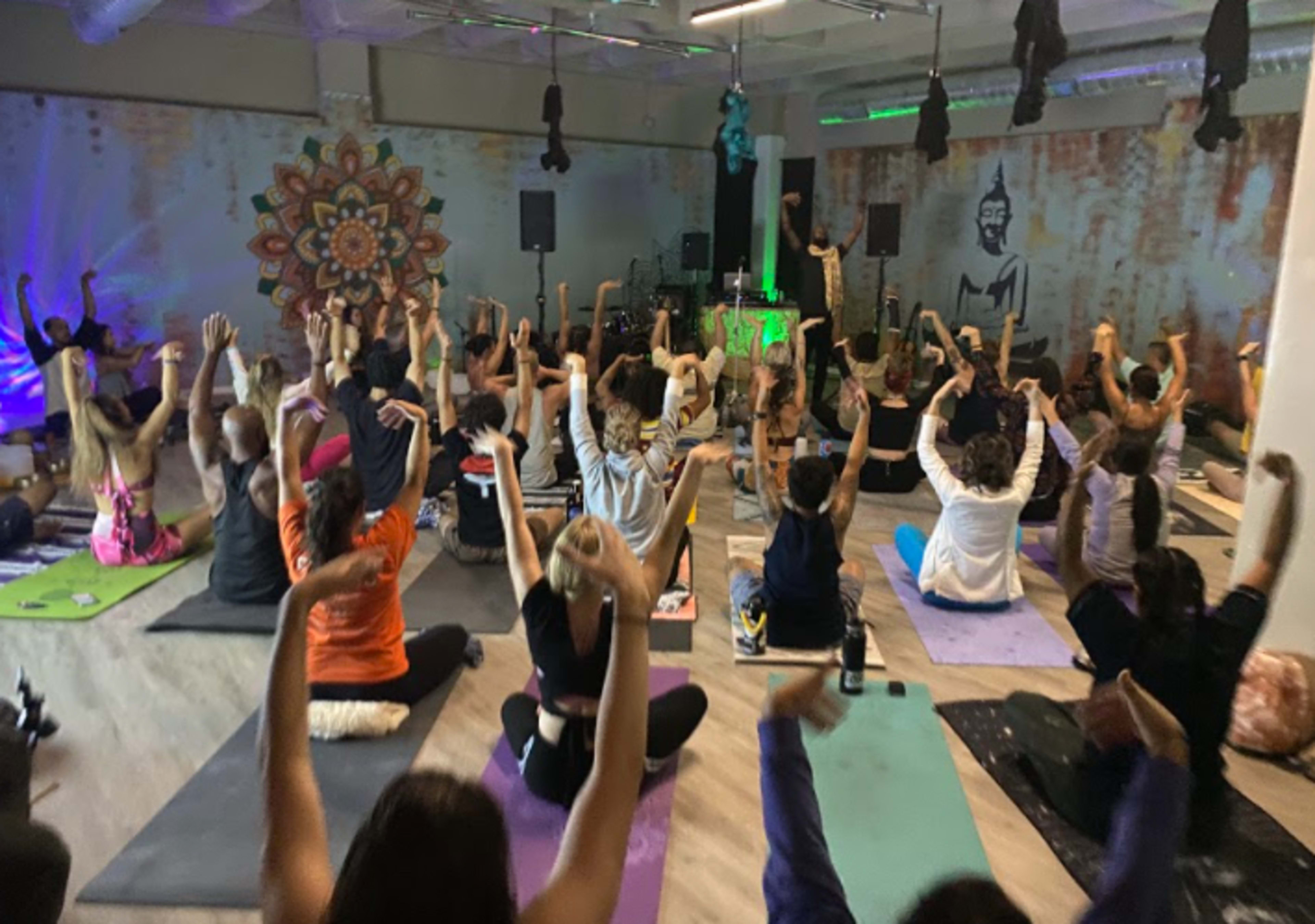 A large group of people participates in a yoga class, raising their arms in a spacious studio decorated with colorful murals and ambient lighting.