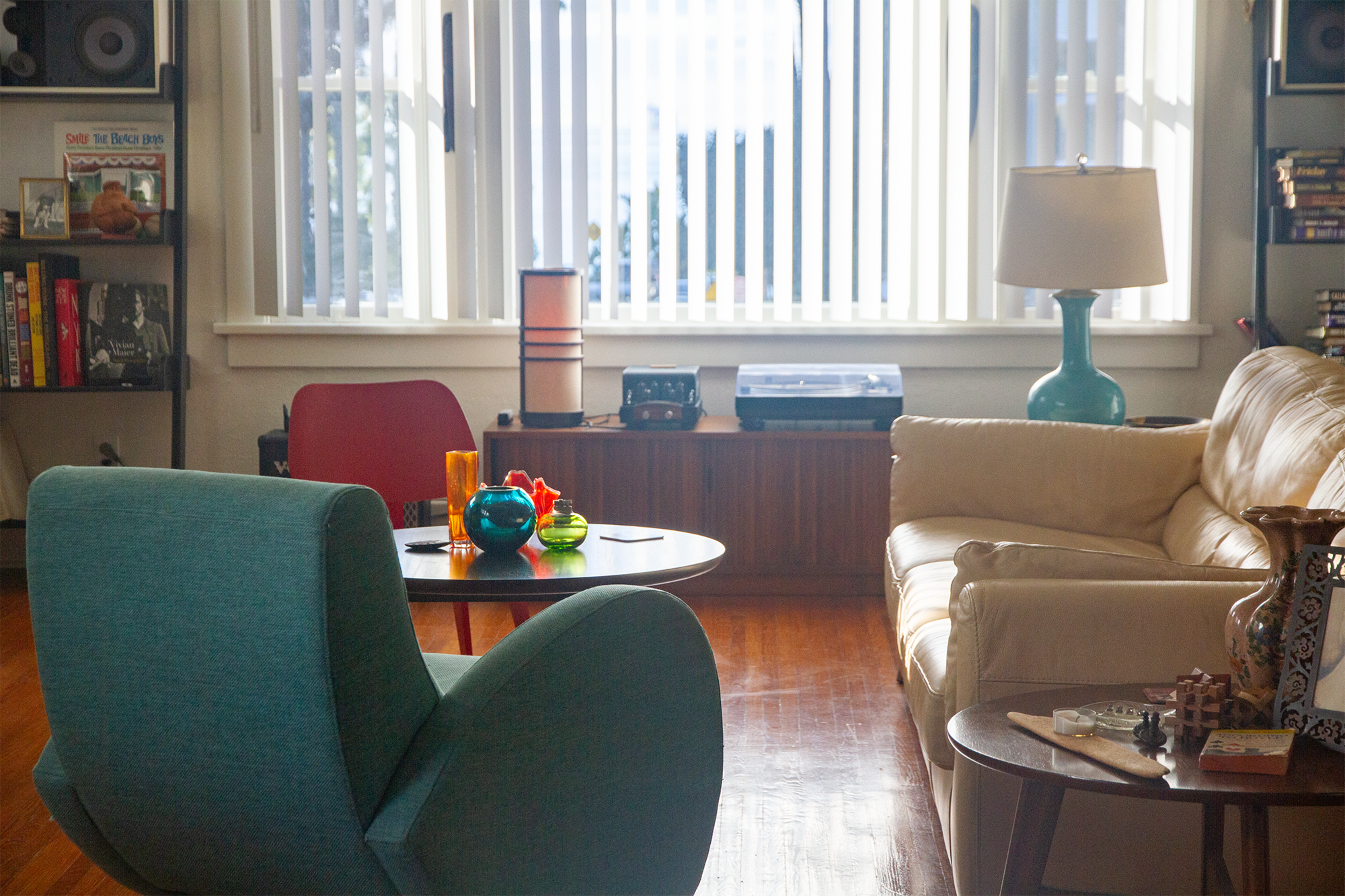 A brightly lit living room features a turquoise chair, a round table with colorful decor, a beige sofa, and a record player beside a large window with vertical blinds.