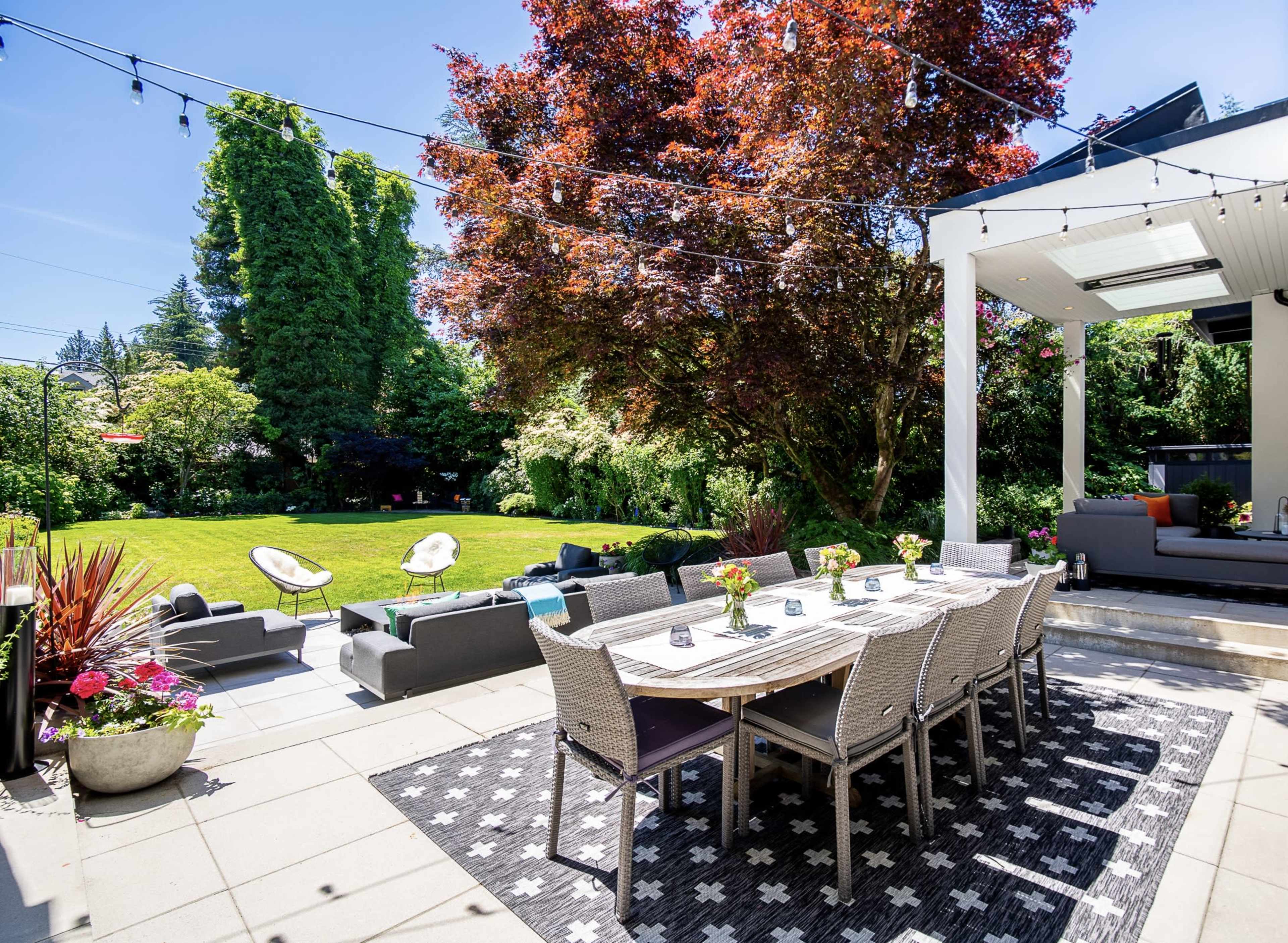 The image shows a patio area with a long dining table set for outdoor meals, surrounded by a well-maintained garden featuring seating and decorative plants.