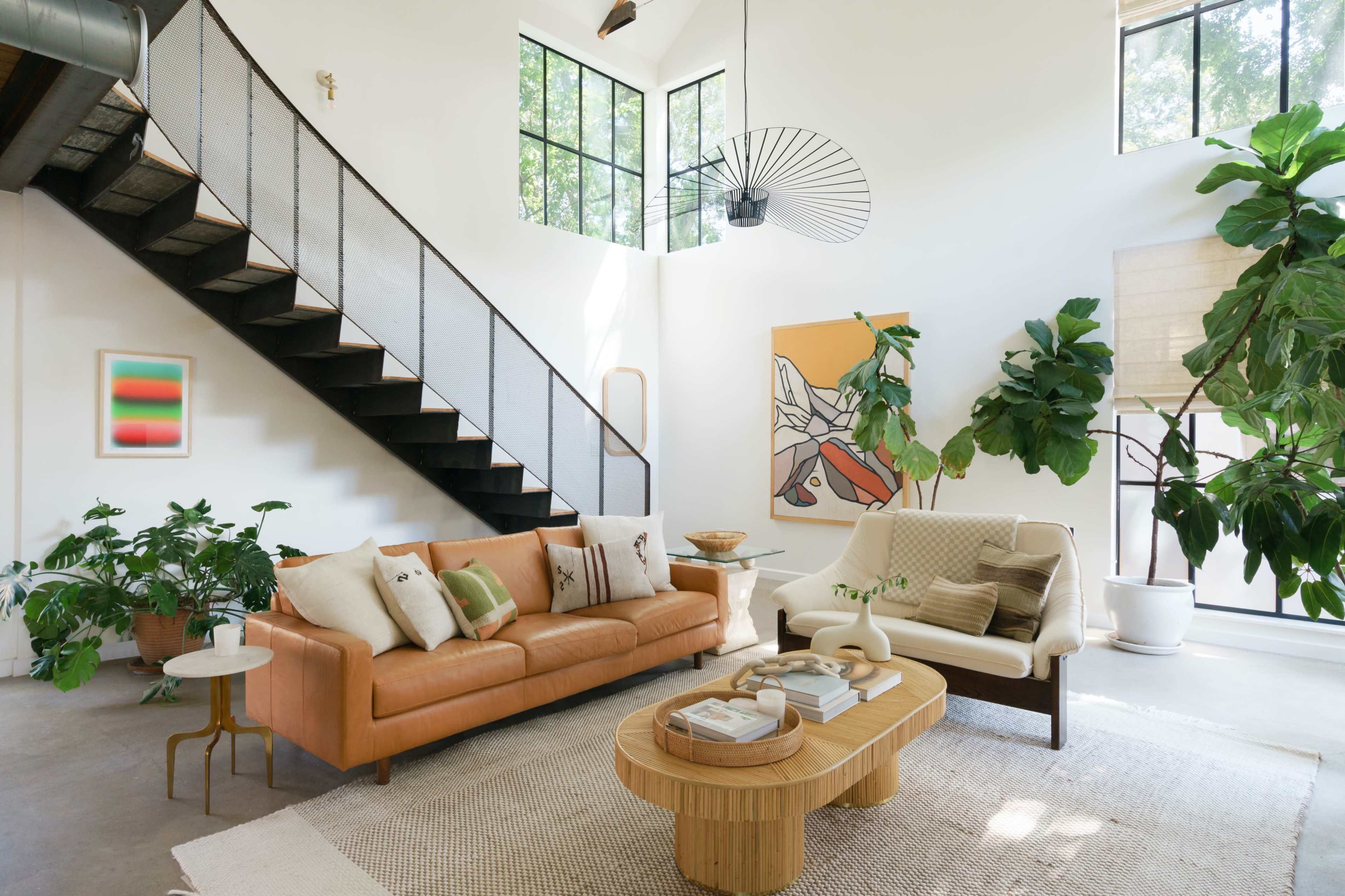 A living room features a staircase, two sofas, a round coffee table, and indoor plants, with large windows allowing natural light to enter.