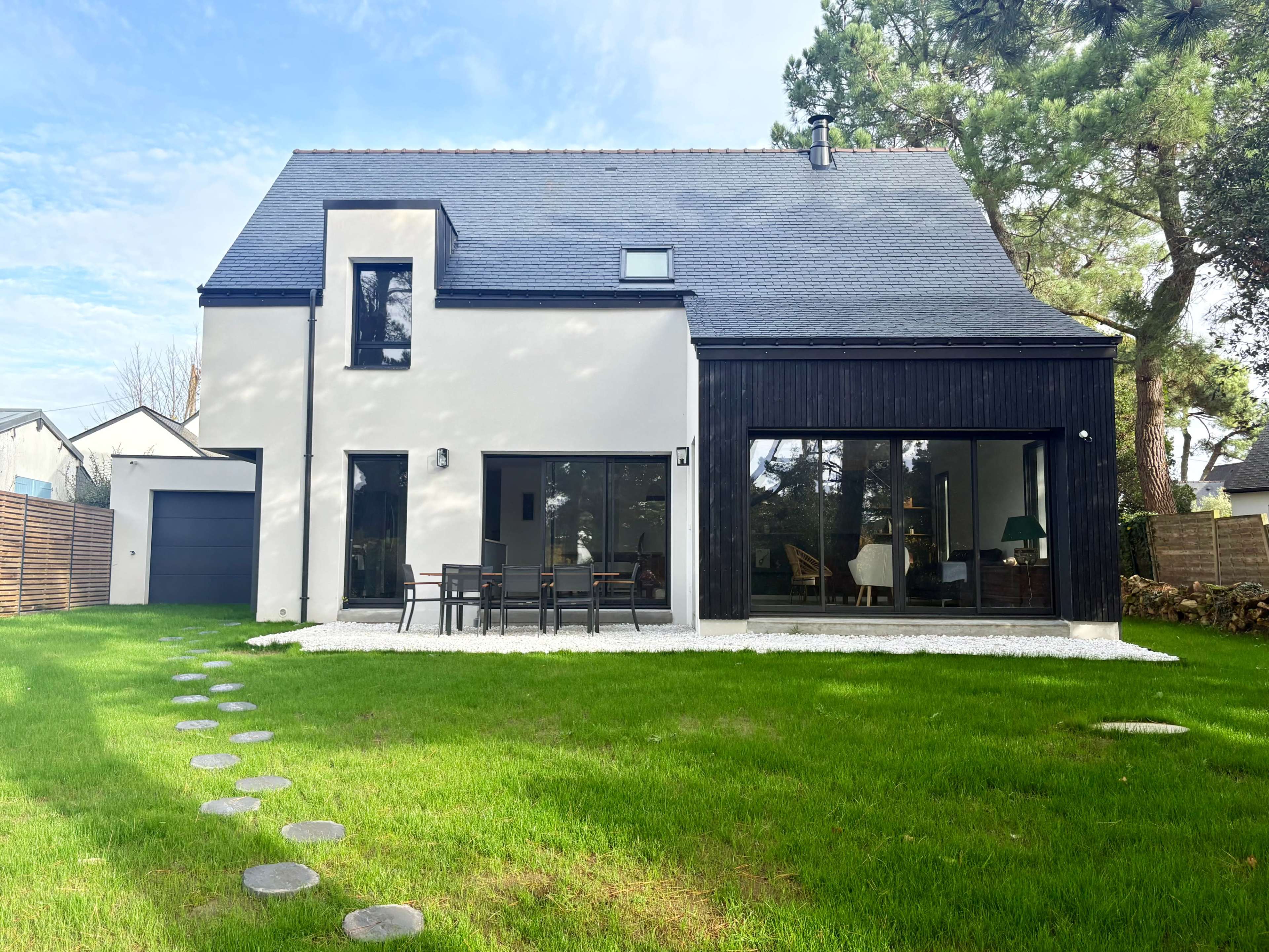 The image shows a modern house with a blend of white walls and a black wooden facade, surrounded by a well-maintained lawn and a stone pathway.