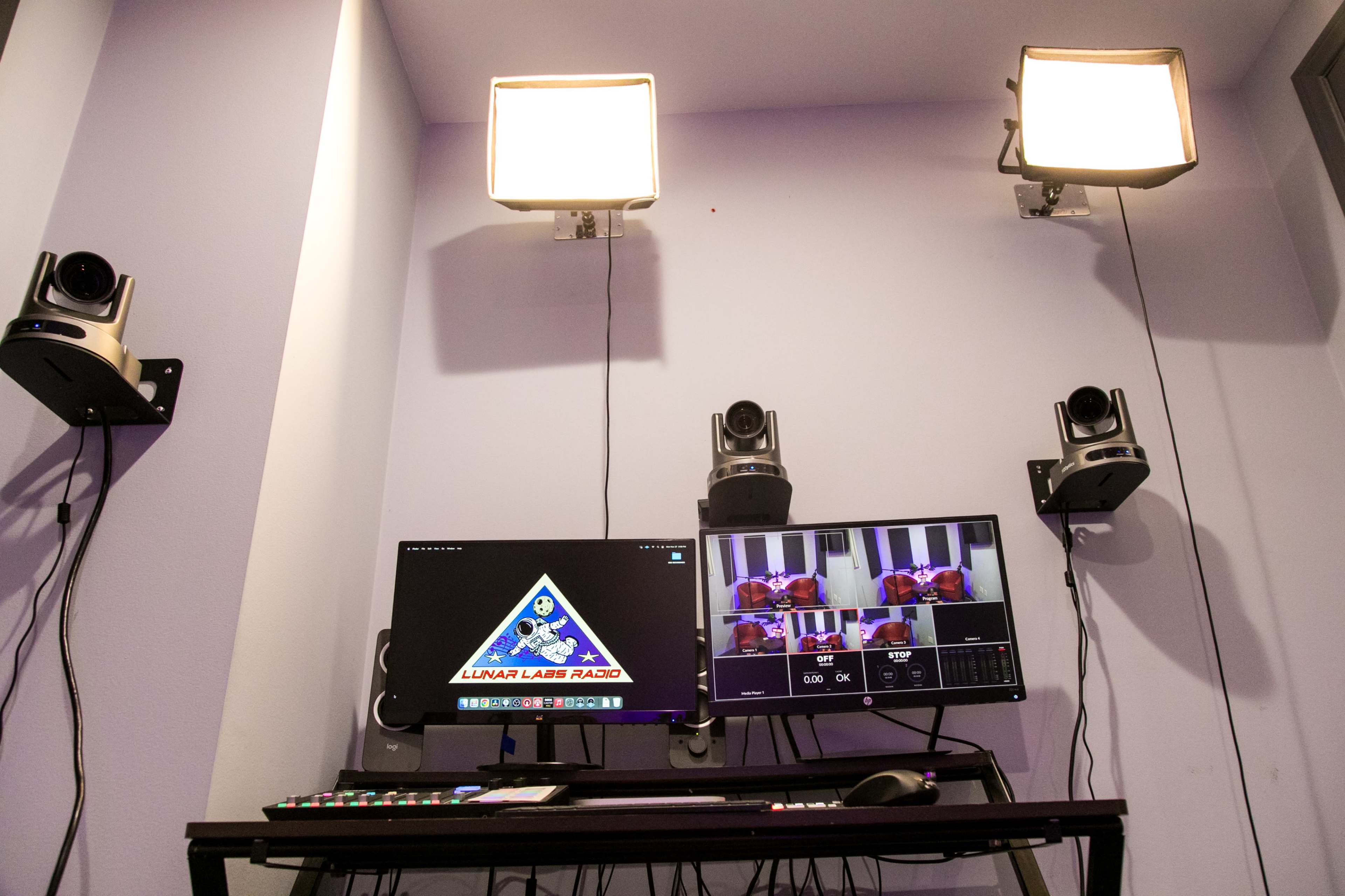 A recording studio with two monitors, overhead lights, and mounted cameras on the walls.