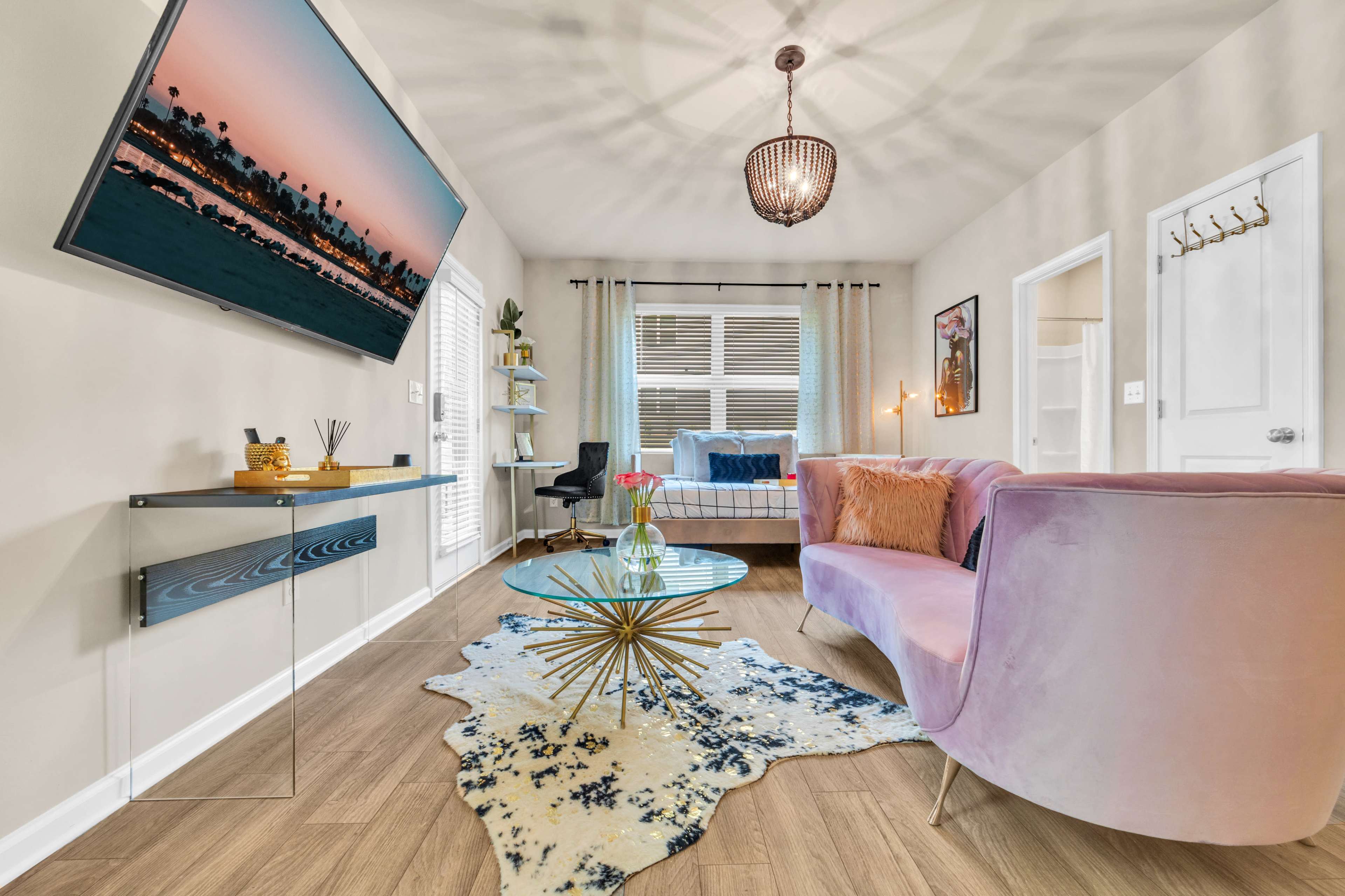 A modern living room features a pink sofa, a round glass coffee table, and a television mounted on the wall, with a decorative rug and natural light coming through the windows.