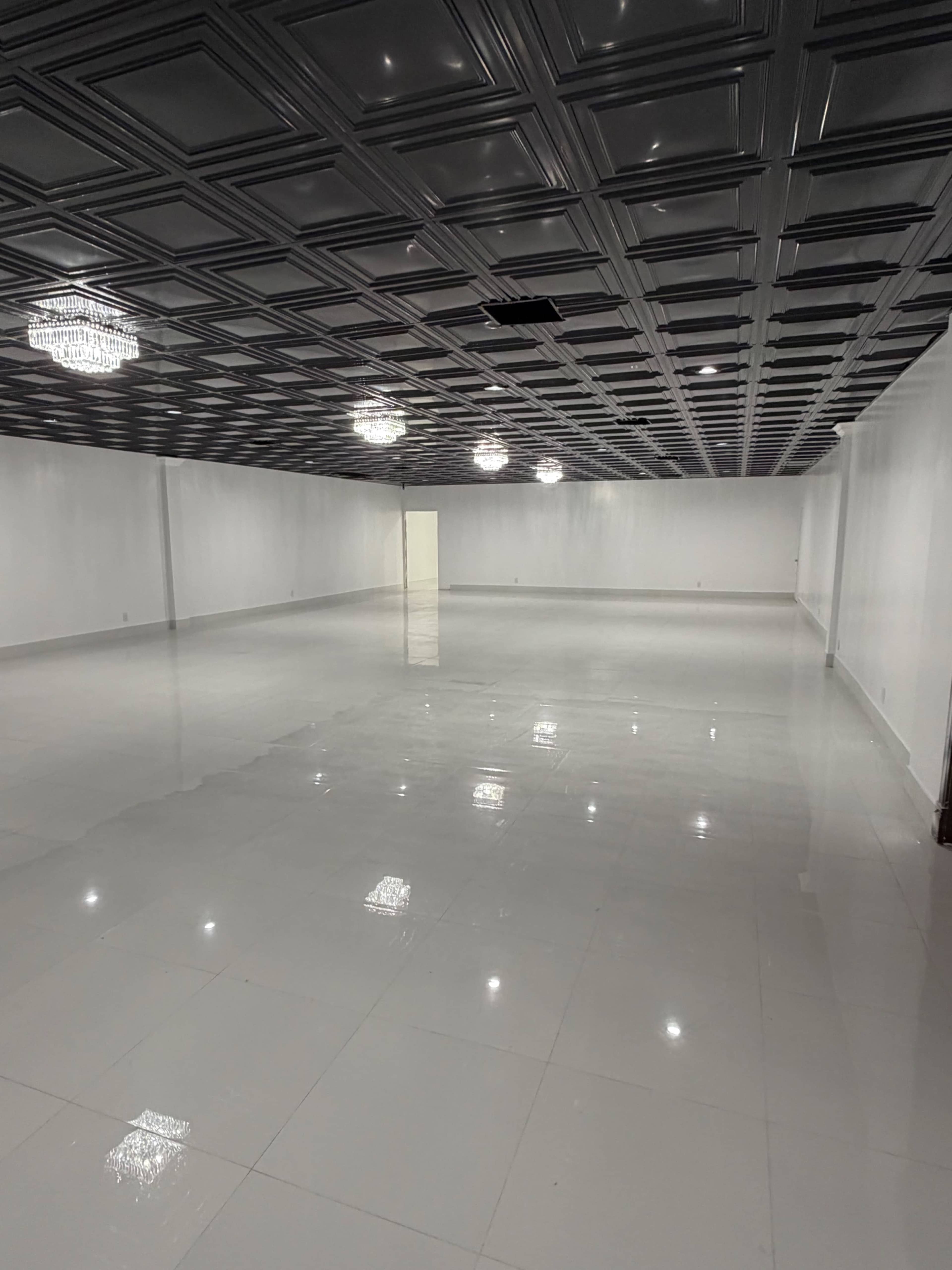 The image shows a large, empty room with a glossy white floor and a ceiling featuring decorative patterns, illuminated by several ceiling lights.