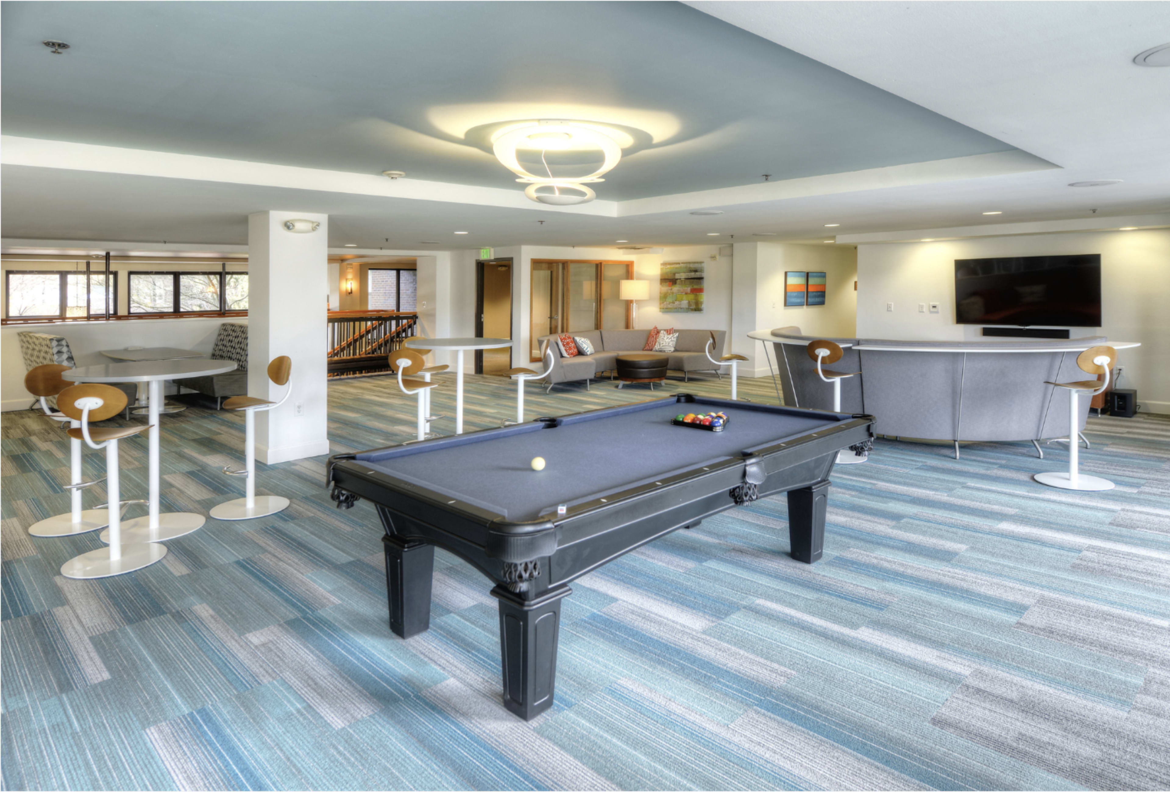 The scene features a modern lounge area with a billiards table in the center, surrounded by seating and a dining space with tall chairs.