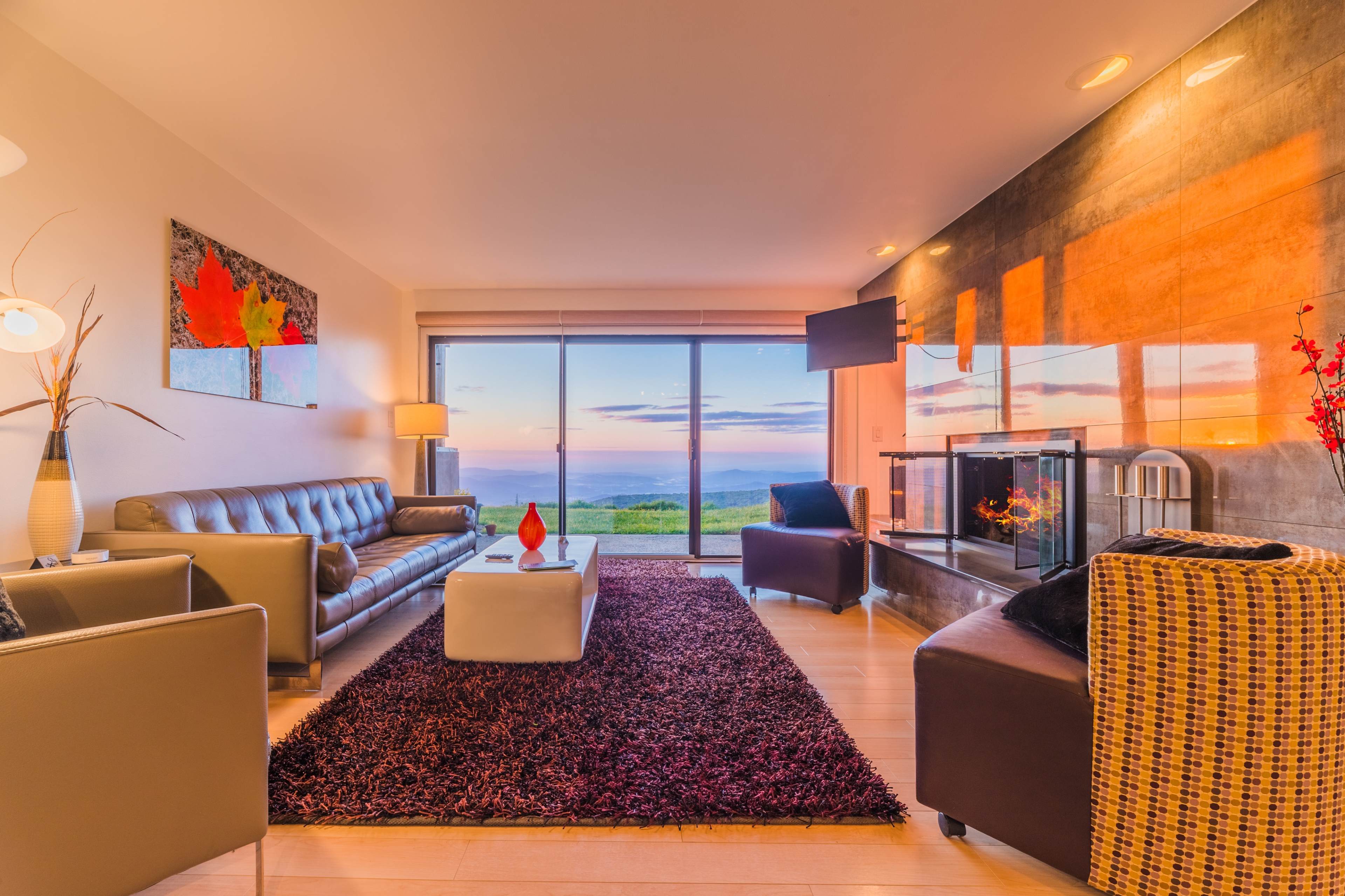 A modern living room with a large window displaying a scenic view and a fireplace, featuring brown leather sofas, a white coffee table, and a plush rug.
