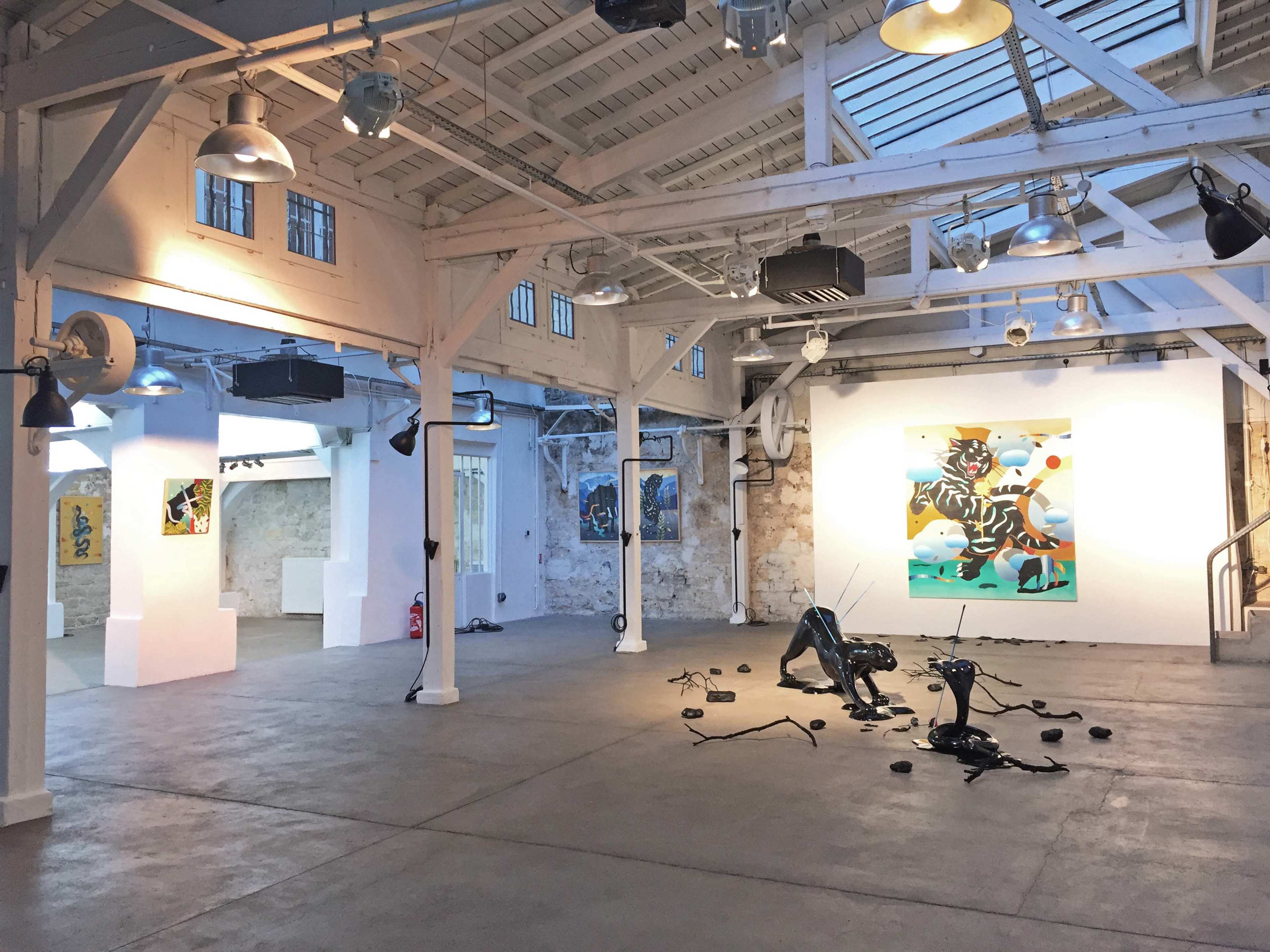 A bright art gallery featuring white walls, exposed beams, and scattered contemporary art pieces, including a large painting and various sculptural installations on the floor.