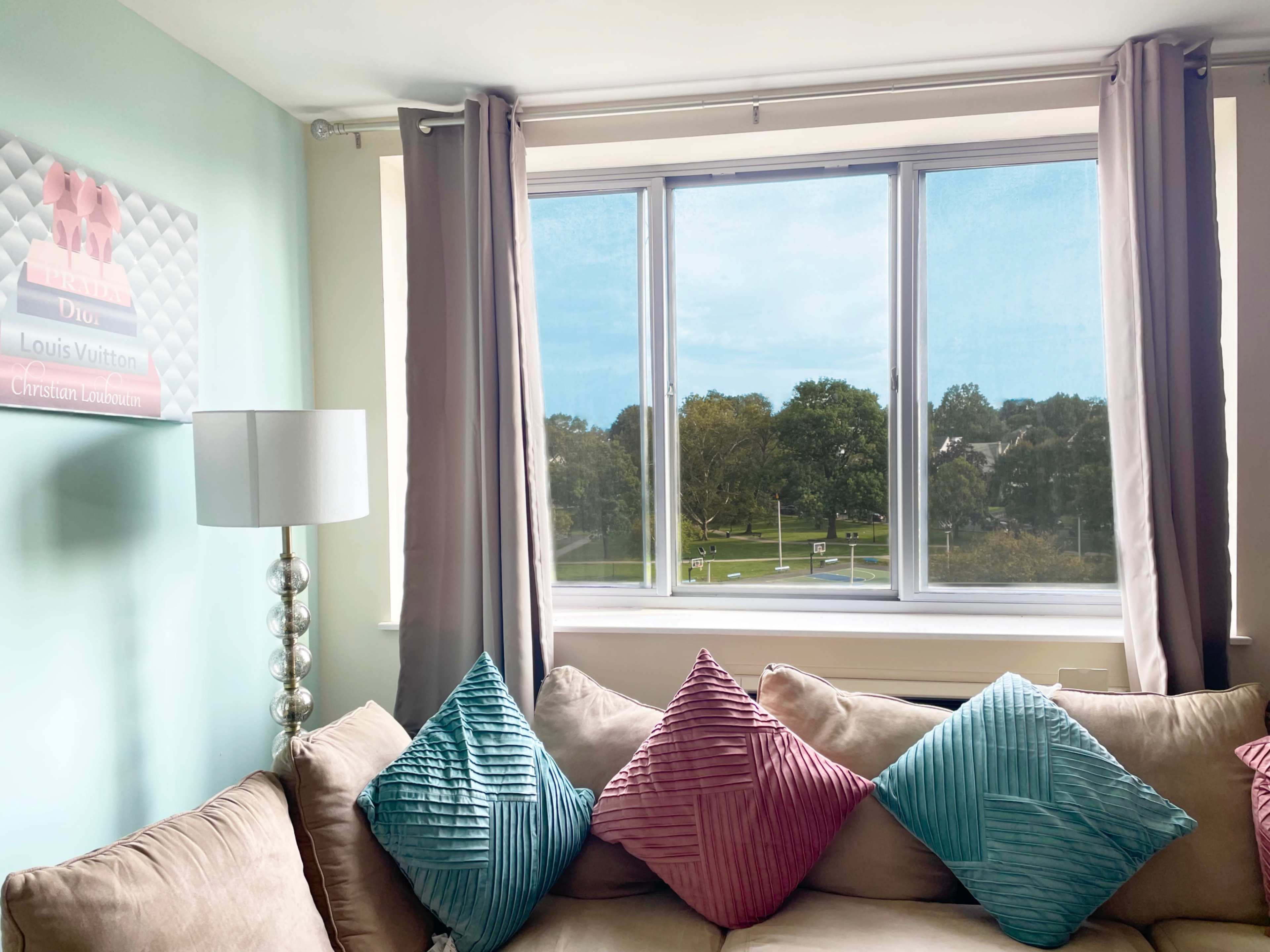 A living room features a beige couch with colorful cushions and a large window overlooking a green landscape.