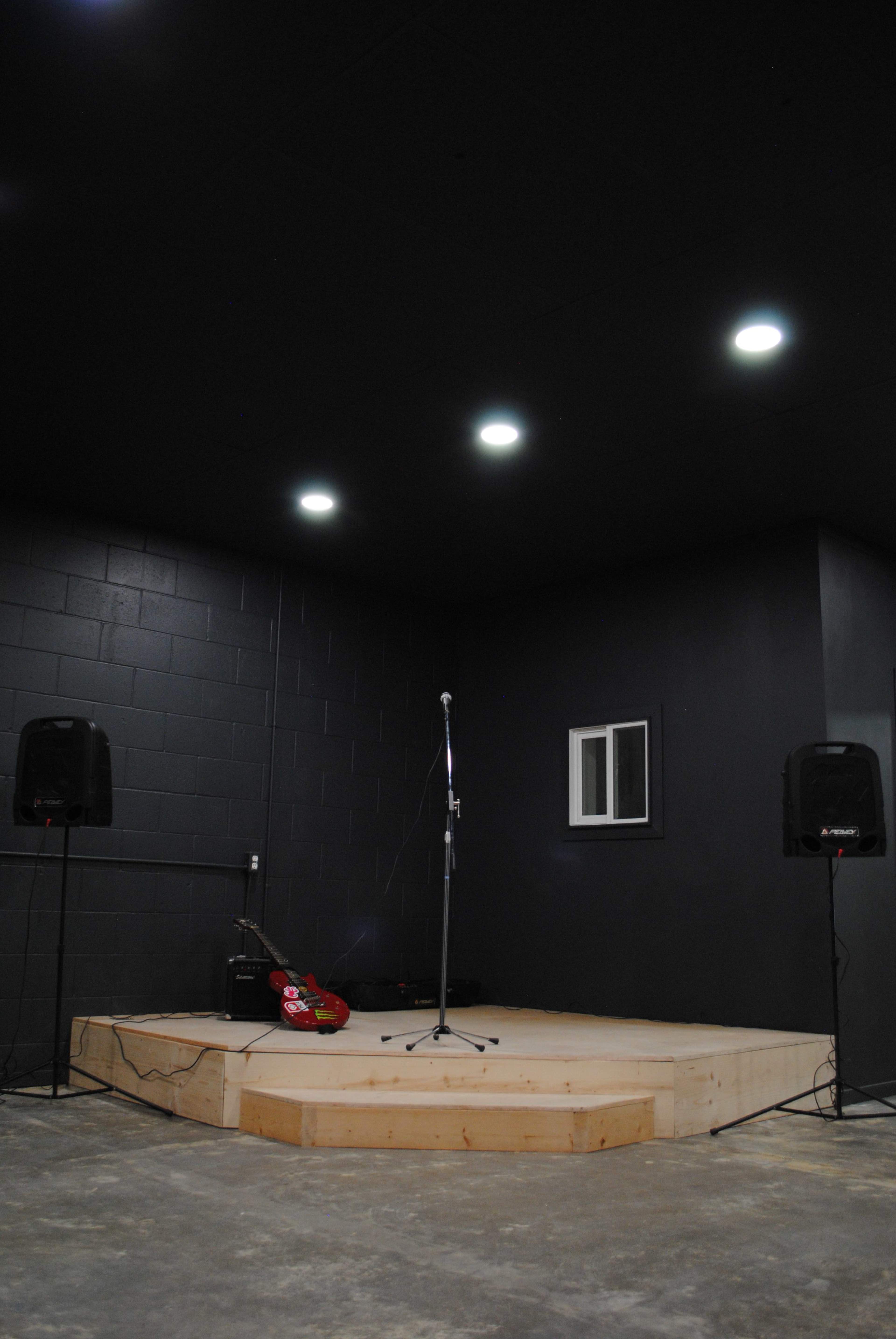 The image shows a small stage with a wooden platform, a microphone stand, two speakers, and a guitar leaning against the wall in a dimly lit room.