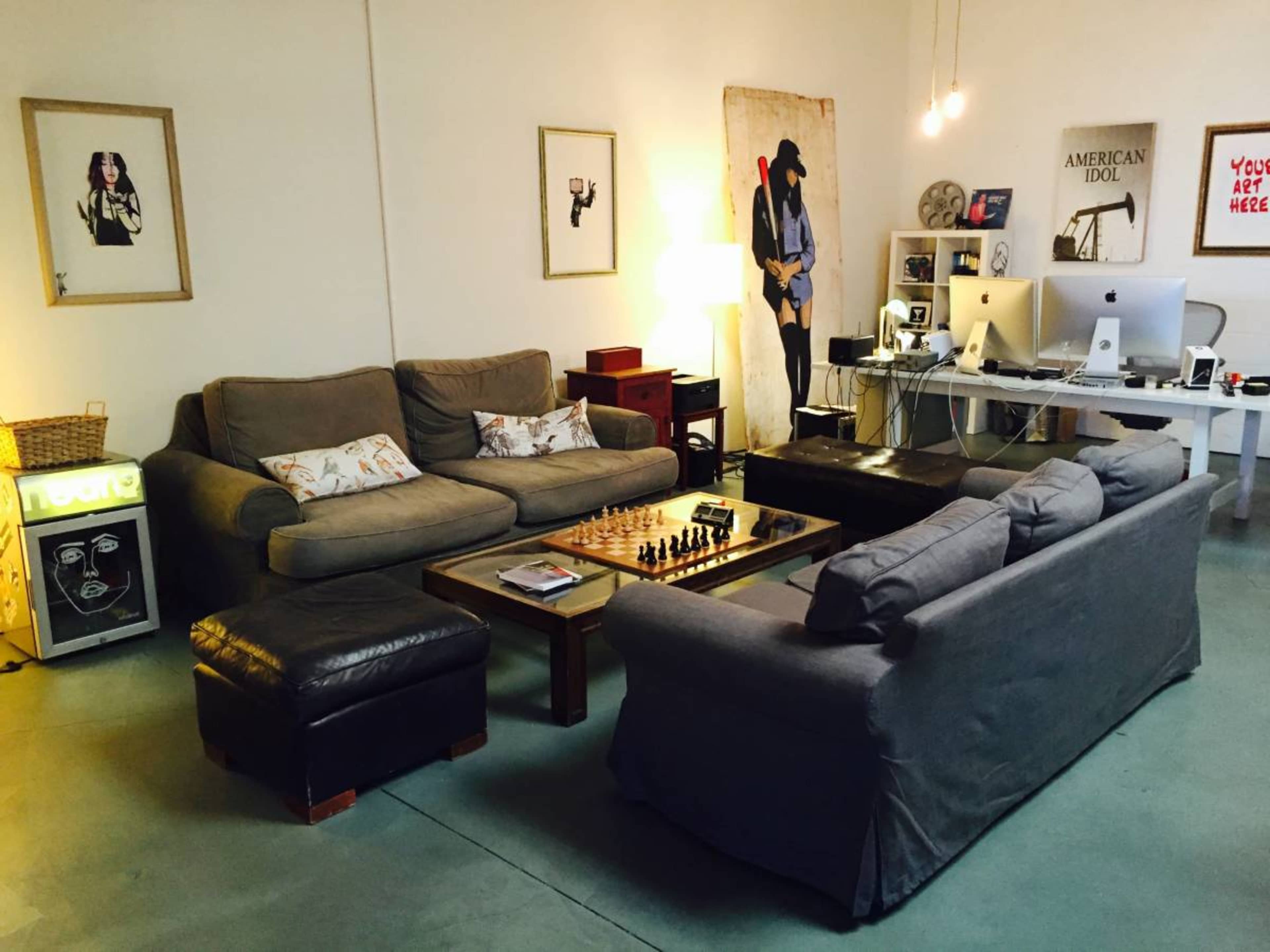 A modern living room with two sofas, a coffee table featuring a chess set, and a work desk with computers and art on the walls.
