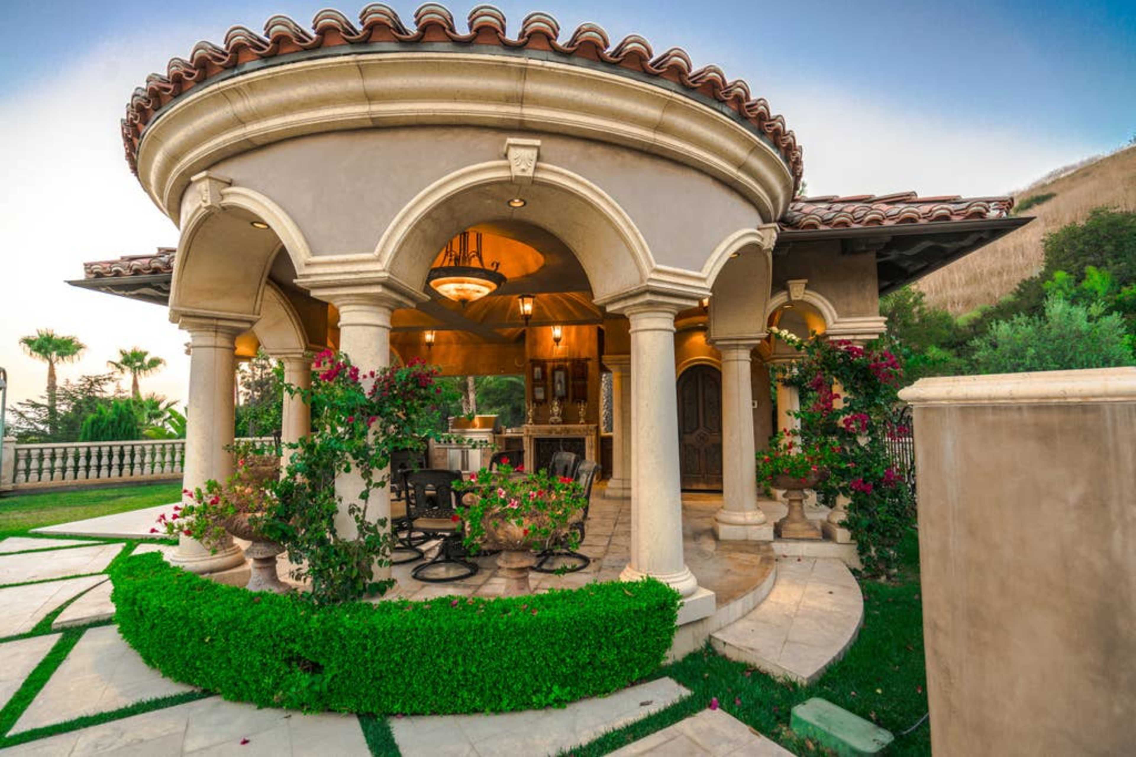 The image shows a beautifully designed round patio with columns, decorated with potted plants and flowers, adjacent to a landscaped lawn.