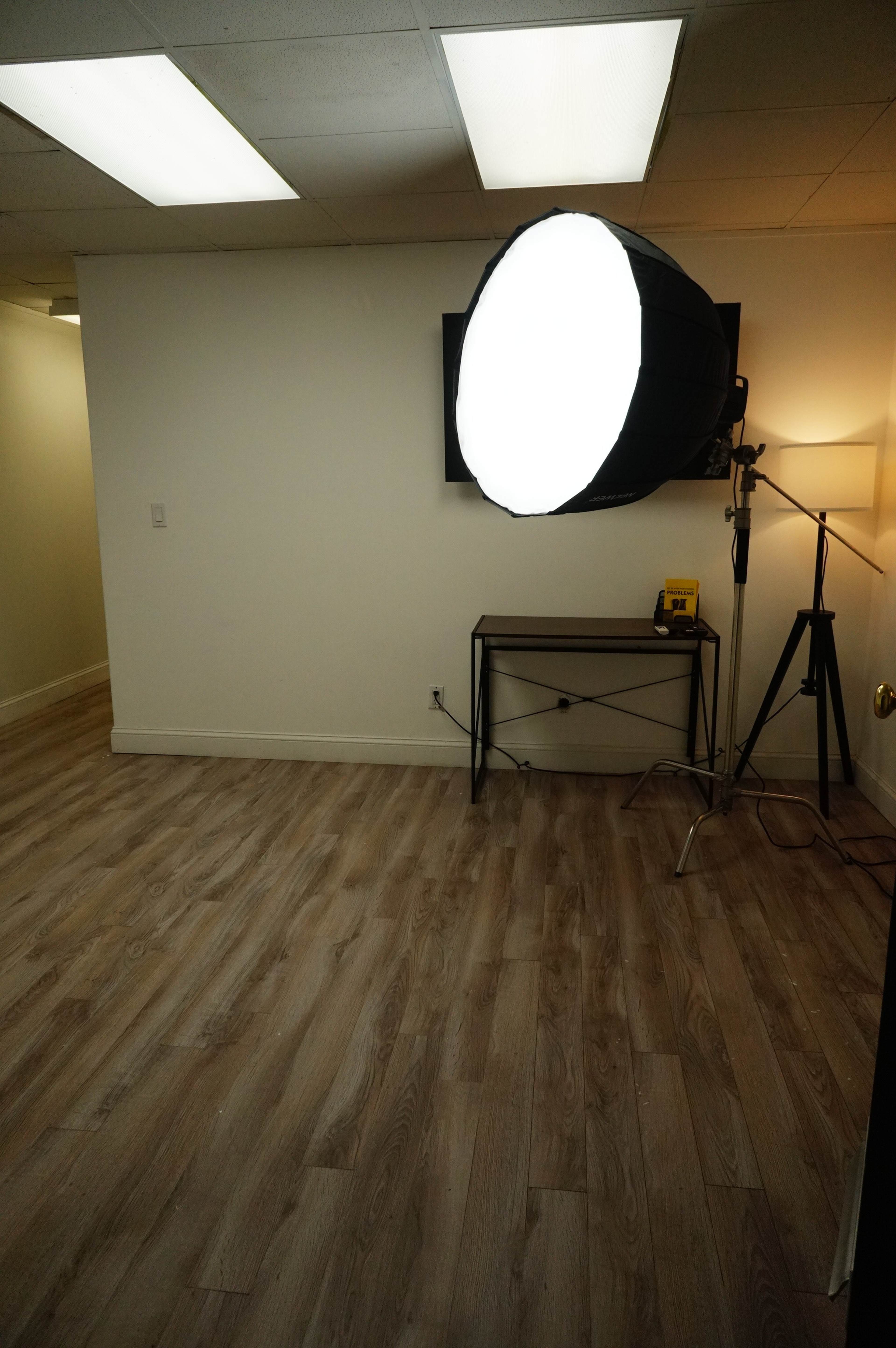 A sparsely furnished room with wooden flooring, a large softbox light on a stand, and a floor lamp in the corner.