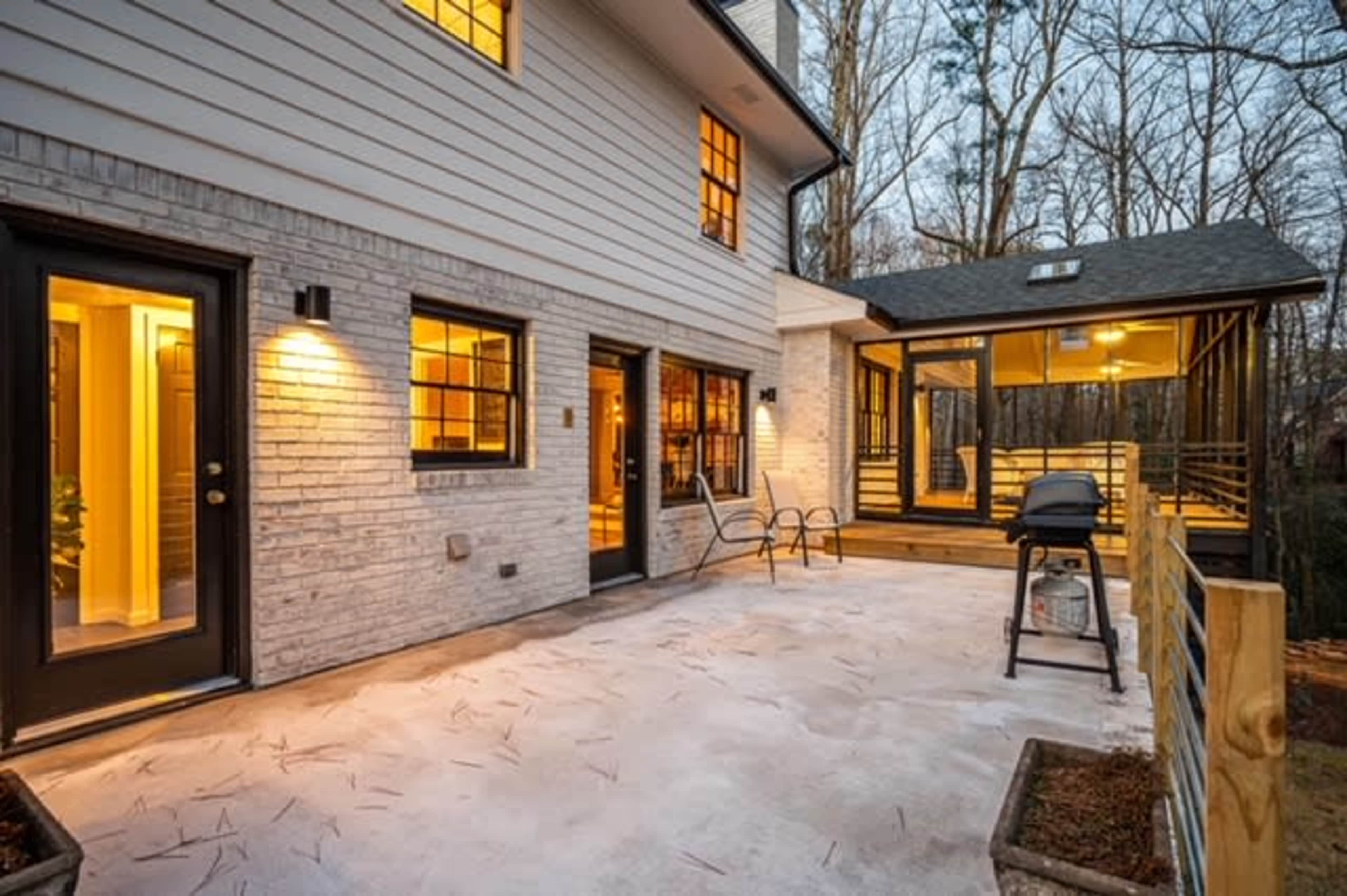 A modern home features a spacious patio area with a grill, surrounded by trees and a screened porch.