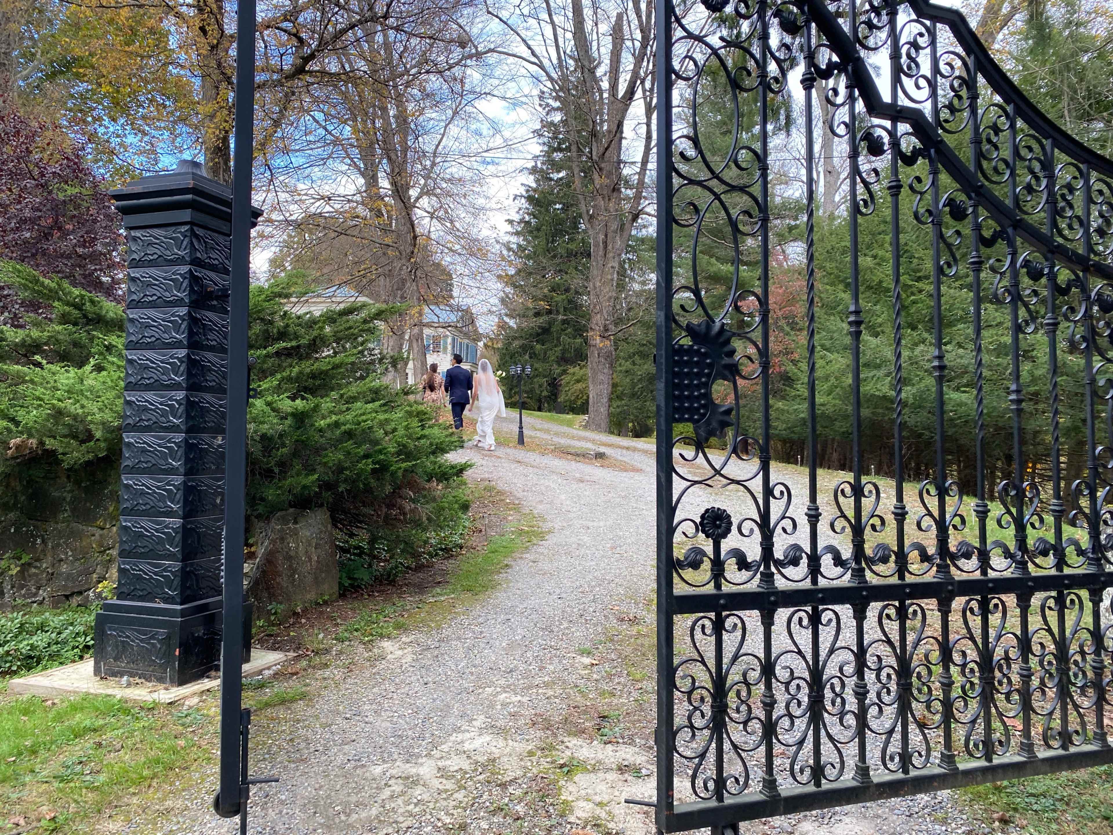 A wrought-iron gate stands open, revealing a gravel path leading to a house in the background where people are walking.