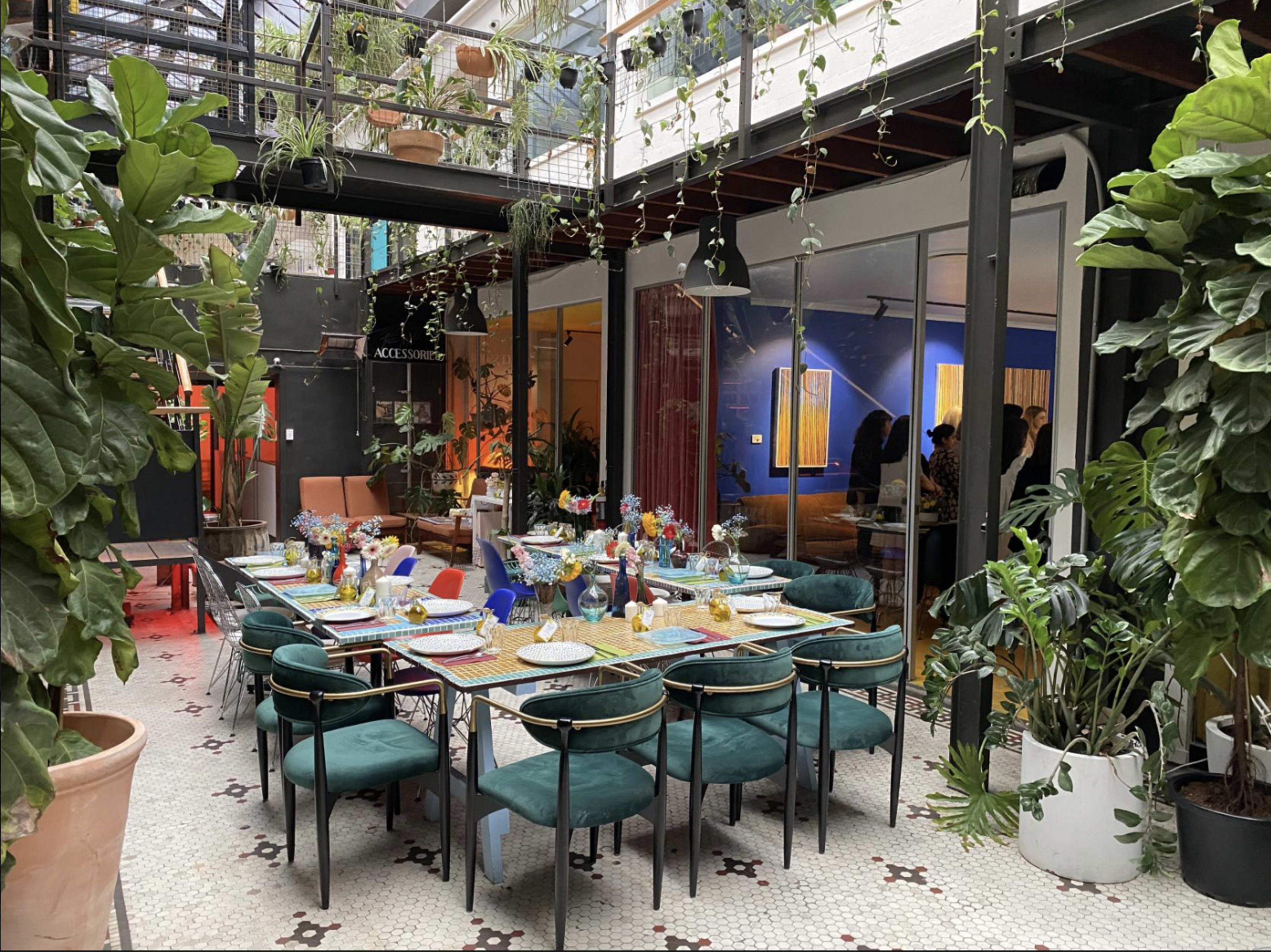 The image shows a dining area set with a long, elegantly arranged table surrounded by green plants and indoor decor.