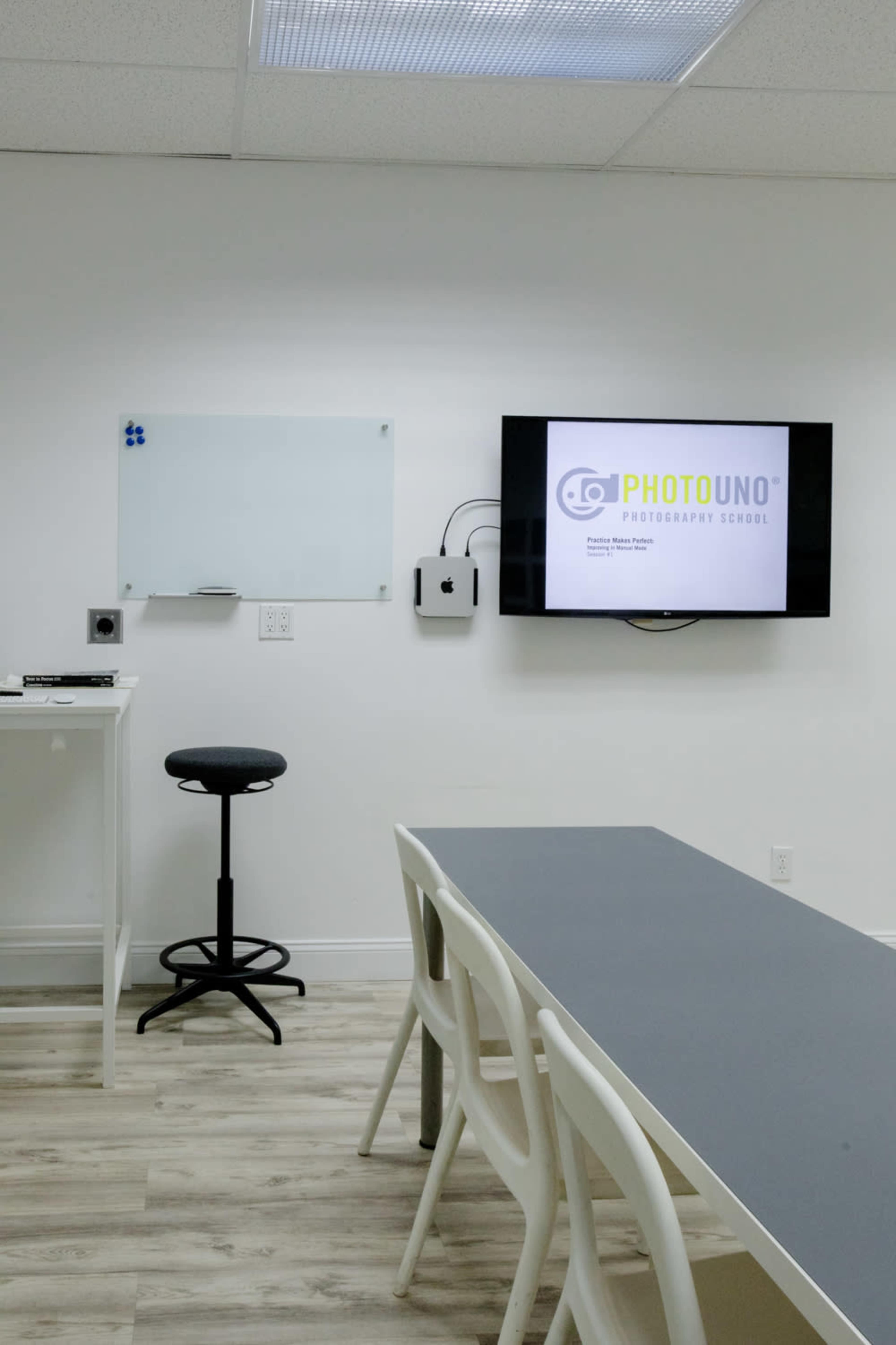 The image shows a minimalist classroom setting with a television displaying "PHOTOUNO Photography School," a whiteboard, a black stool, and a long table.