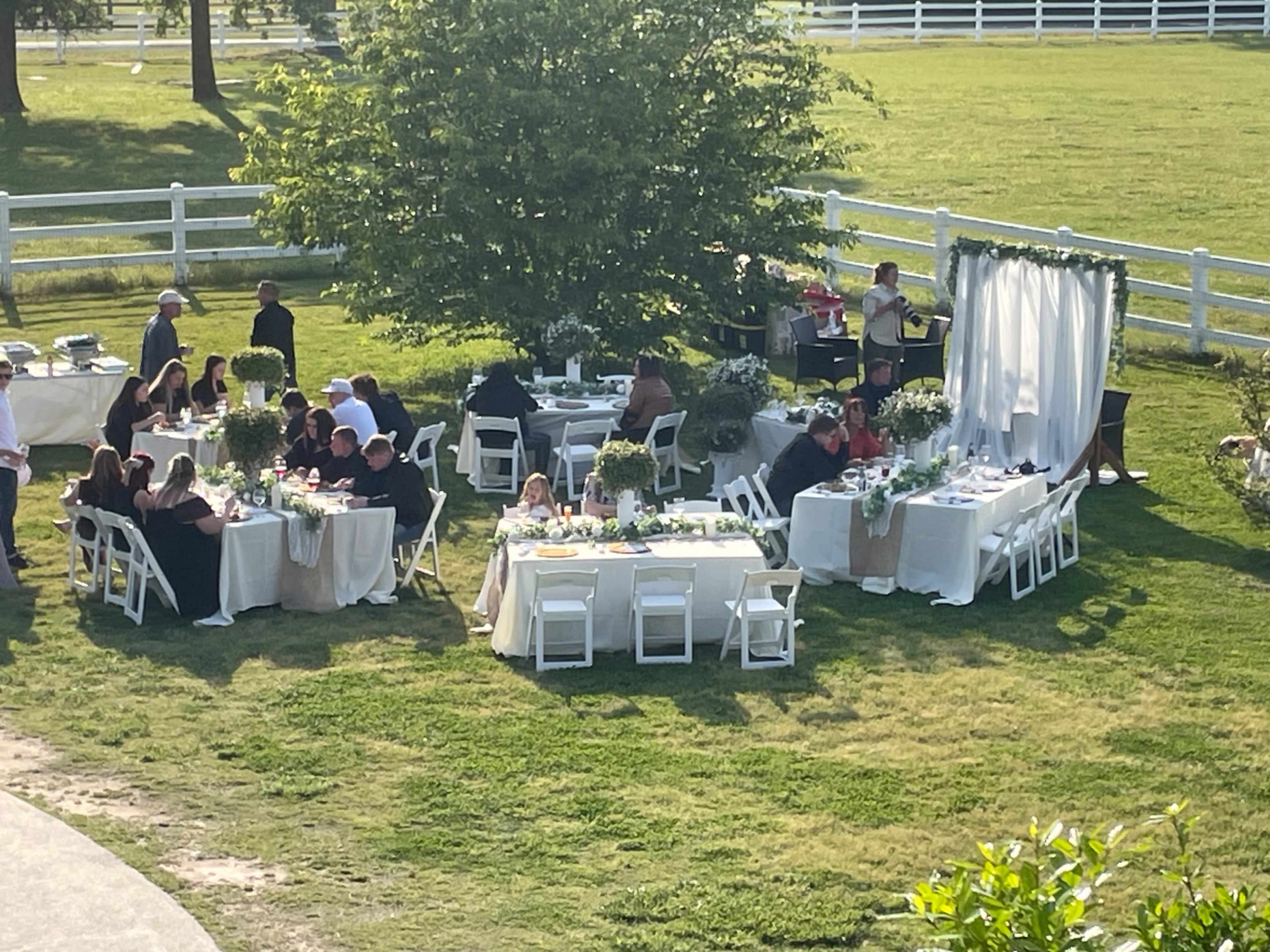 A group of people are seated at multiple tables set up outdoors for a gathering, surrounded by lush greenery and a white fence.