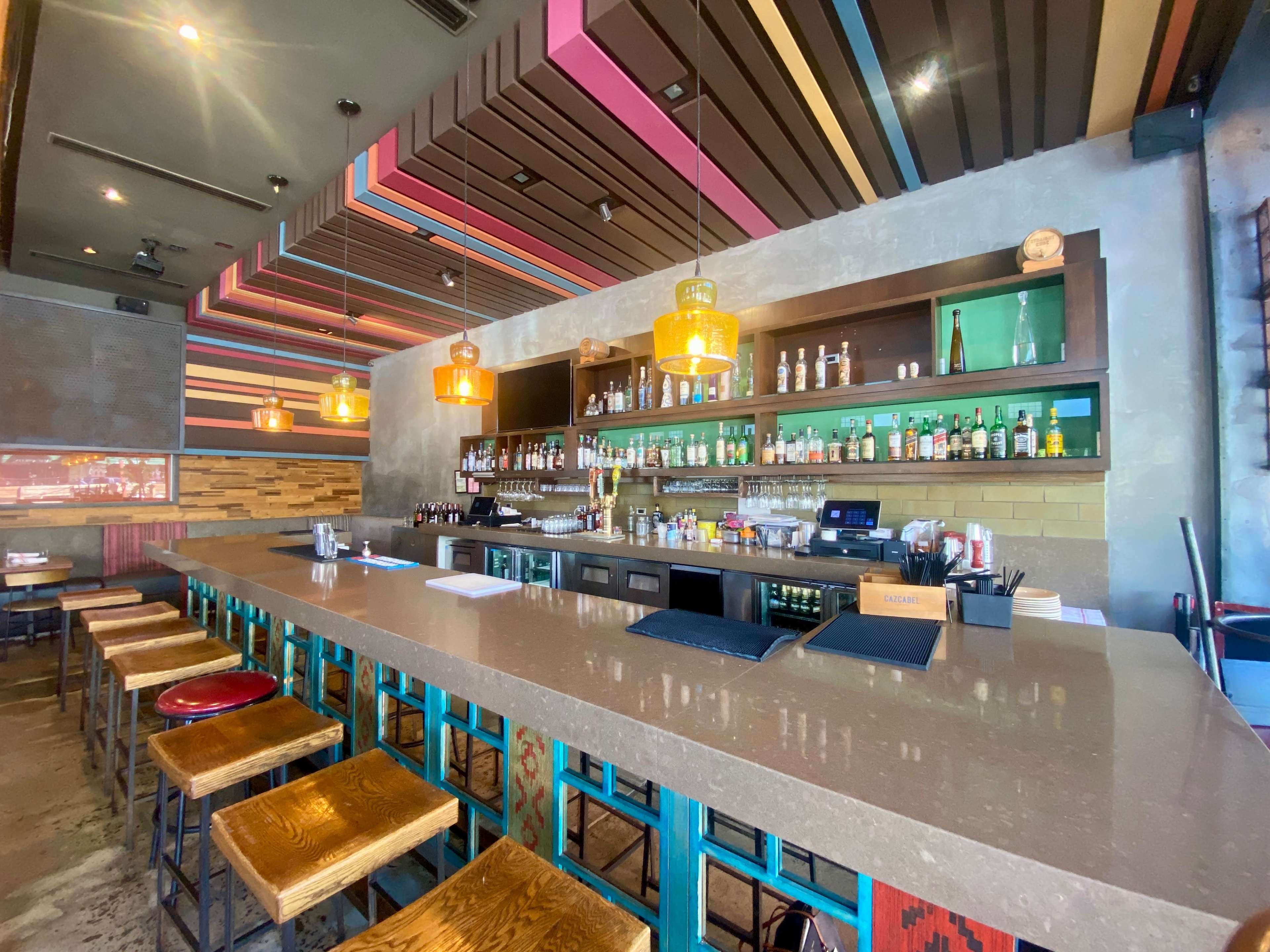 A modern bar with a long countertop, colorful overhead lighting, and shelves stocked with various bottles behind it.