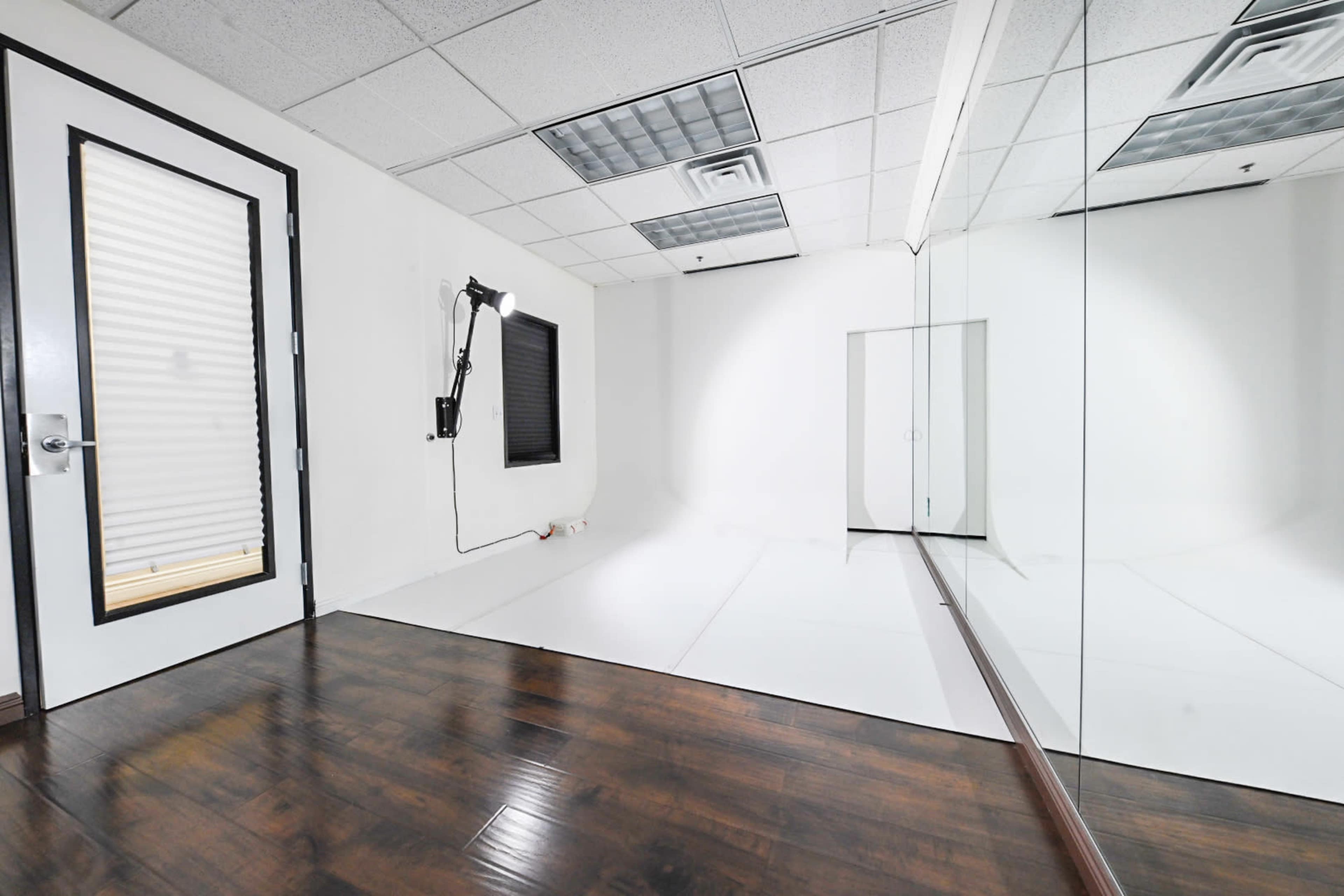 The image shows a minimalist studio space with white walls, a door, a large mirror, and a bright, uncluttered floor.