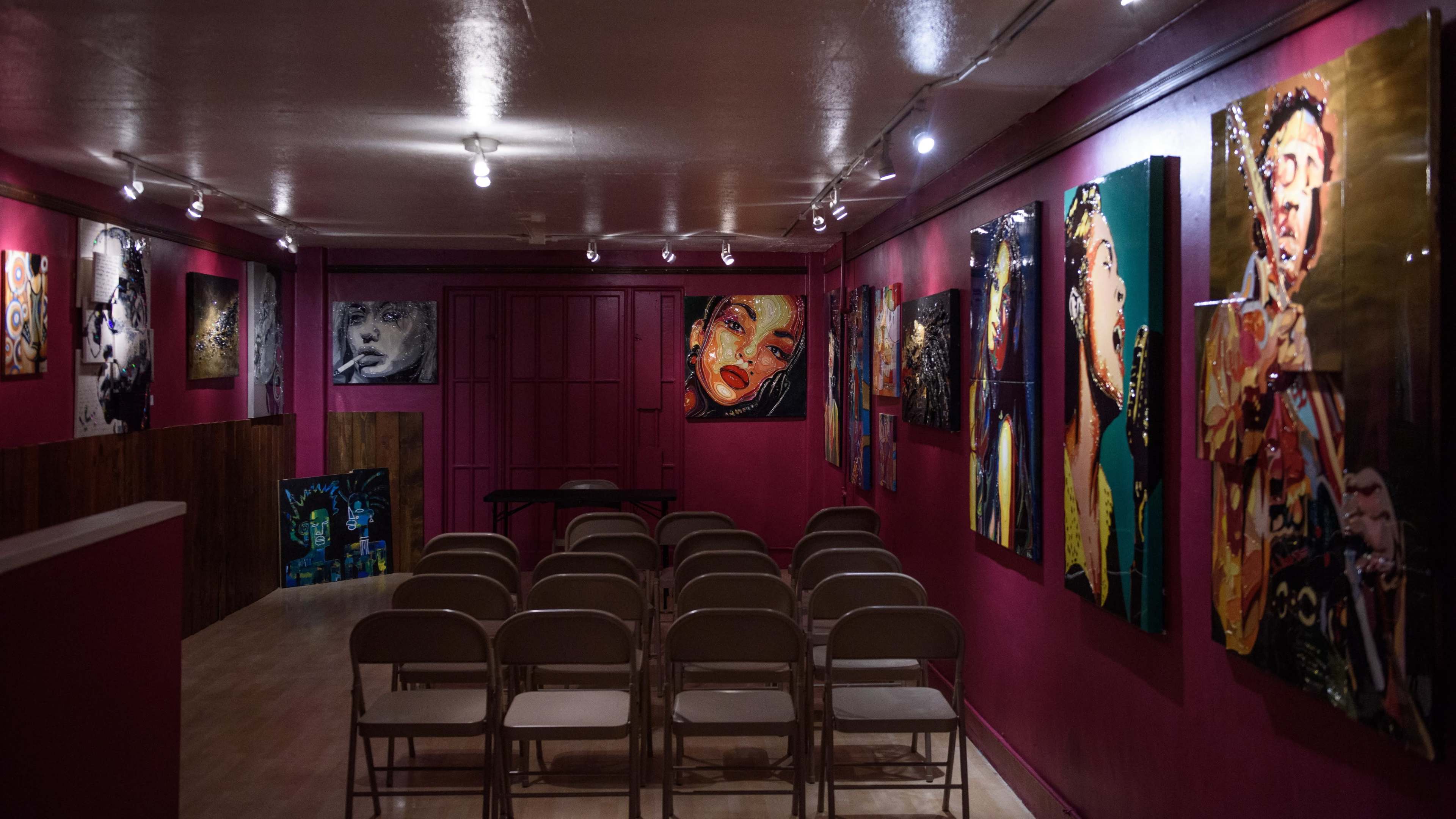 A gallery room with purple walls, featuring several colorful paintings, and rows of folding chairs arranged for an audience.
