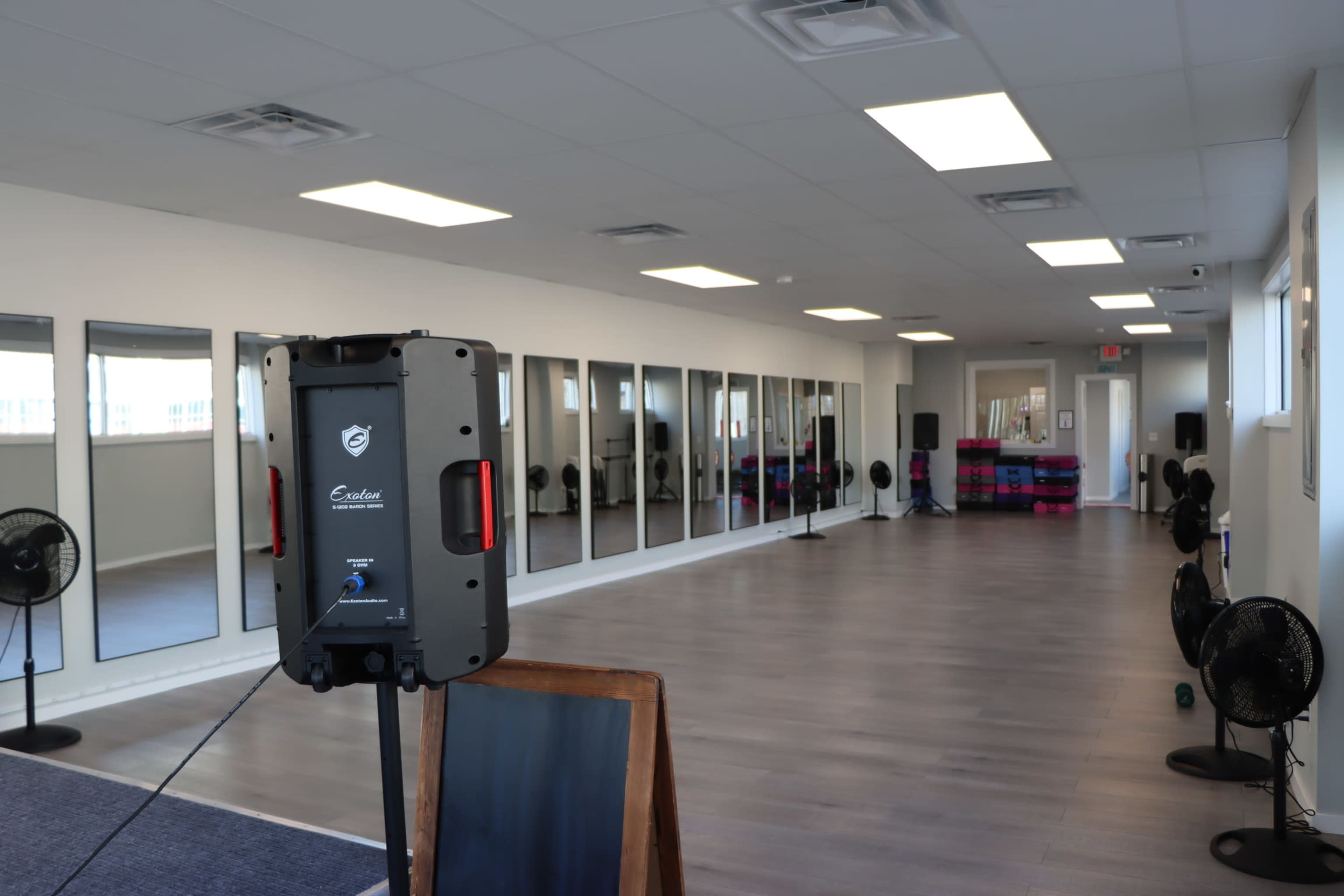 The image shows an empty room with large mirrors lining one wall, wooden flooring, and several fans positioned around the space.