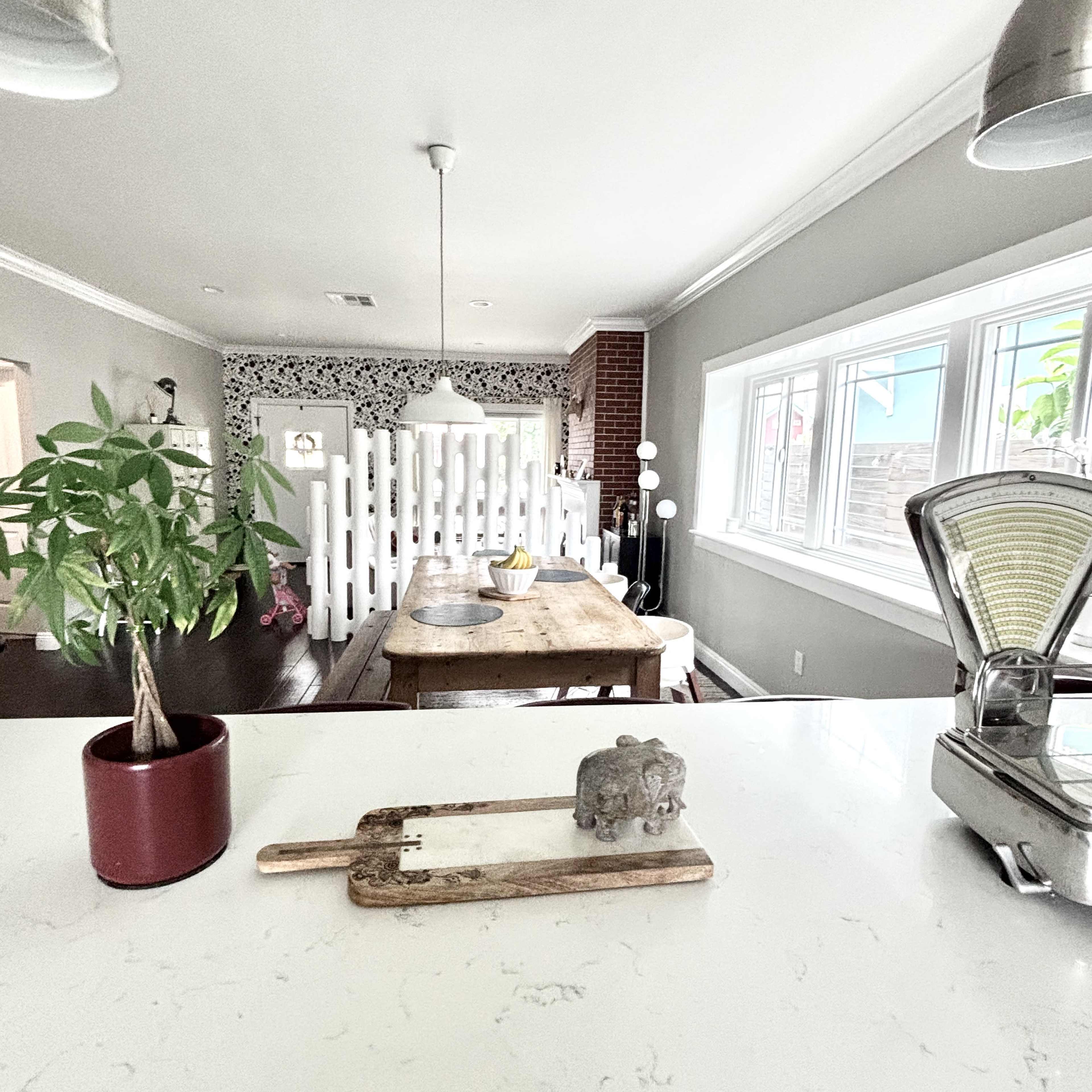 The image shows a kitchen and dining area with a wooden table, a potted plant, and a white partition separating the space from a living area with patterned walls.