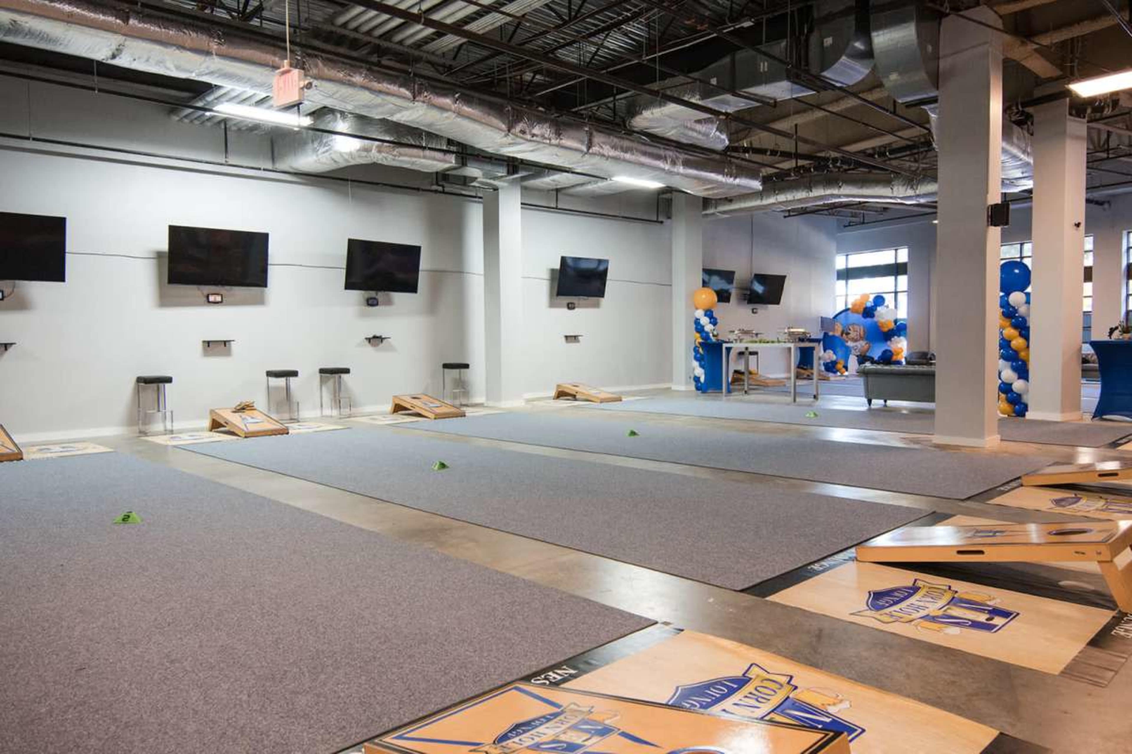 The image shows an indoor recreational area featuring several cornhole boards set up on a carpeted surface, with television screens mounted on the walls and blue-and-yellow balloons in the corner.